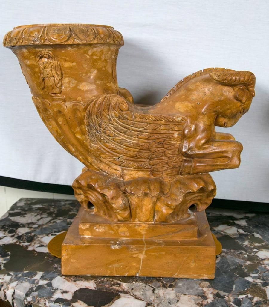 A very interesting and different pair of hand-carved solid marble decorative items. Integral stepped base with shell type carving above. A winged ram, front legs tucked in, sits atop. From the backs rise a trumpet form, decorated with robed figures
