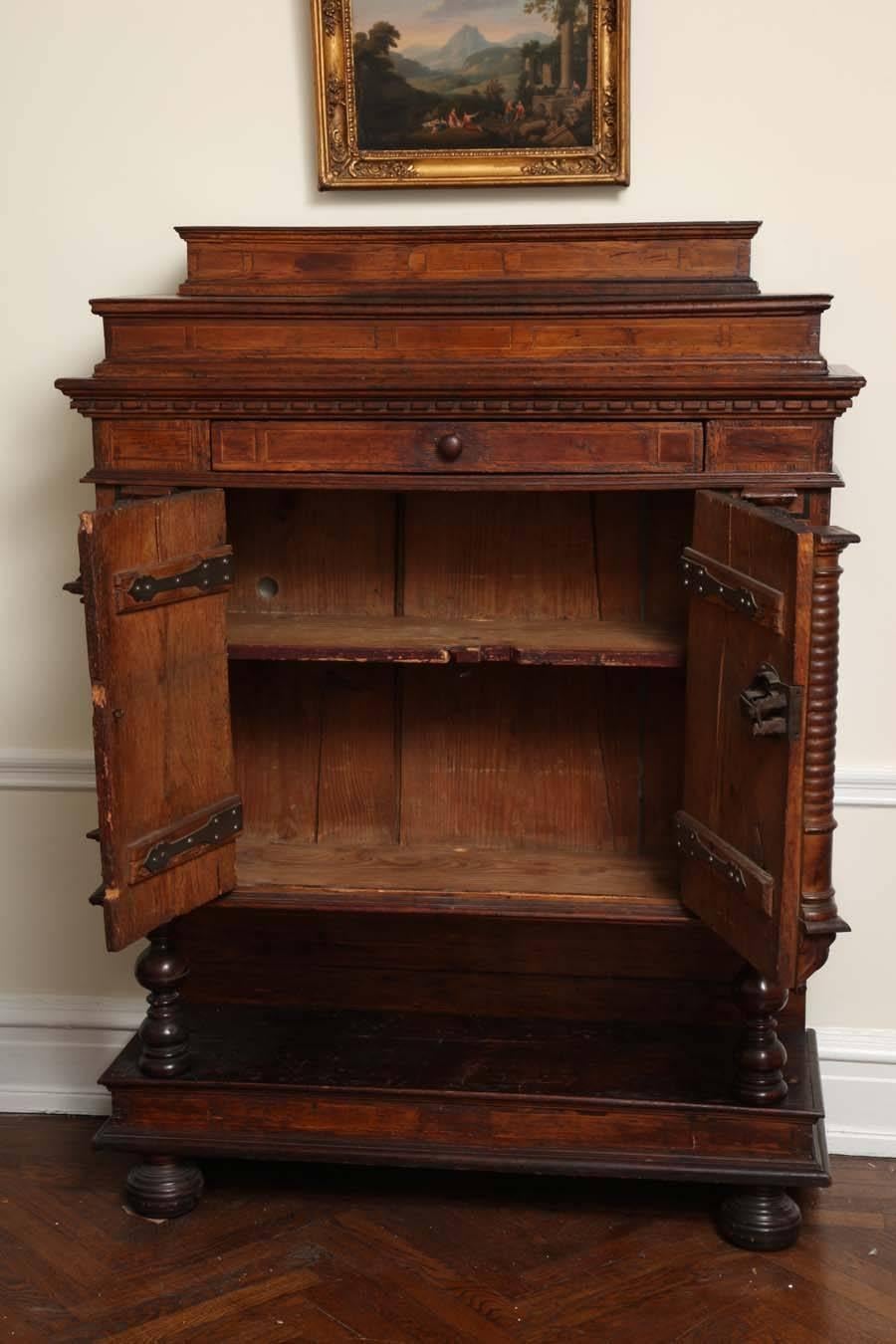 Unusual cabinet surmounted by graduated tiers with geometric marquetry, over a moulded cornice and frieze drawer over two doors elaborately carved with putti and classical masks set between turn engaged columns; raised on turned supports on a