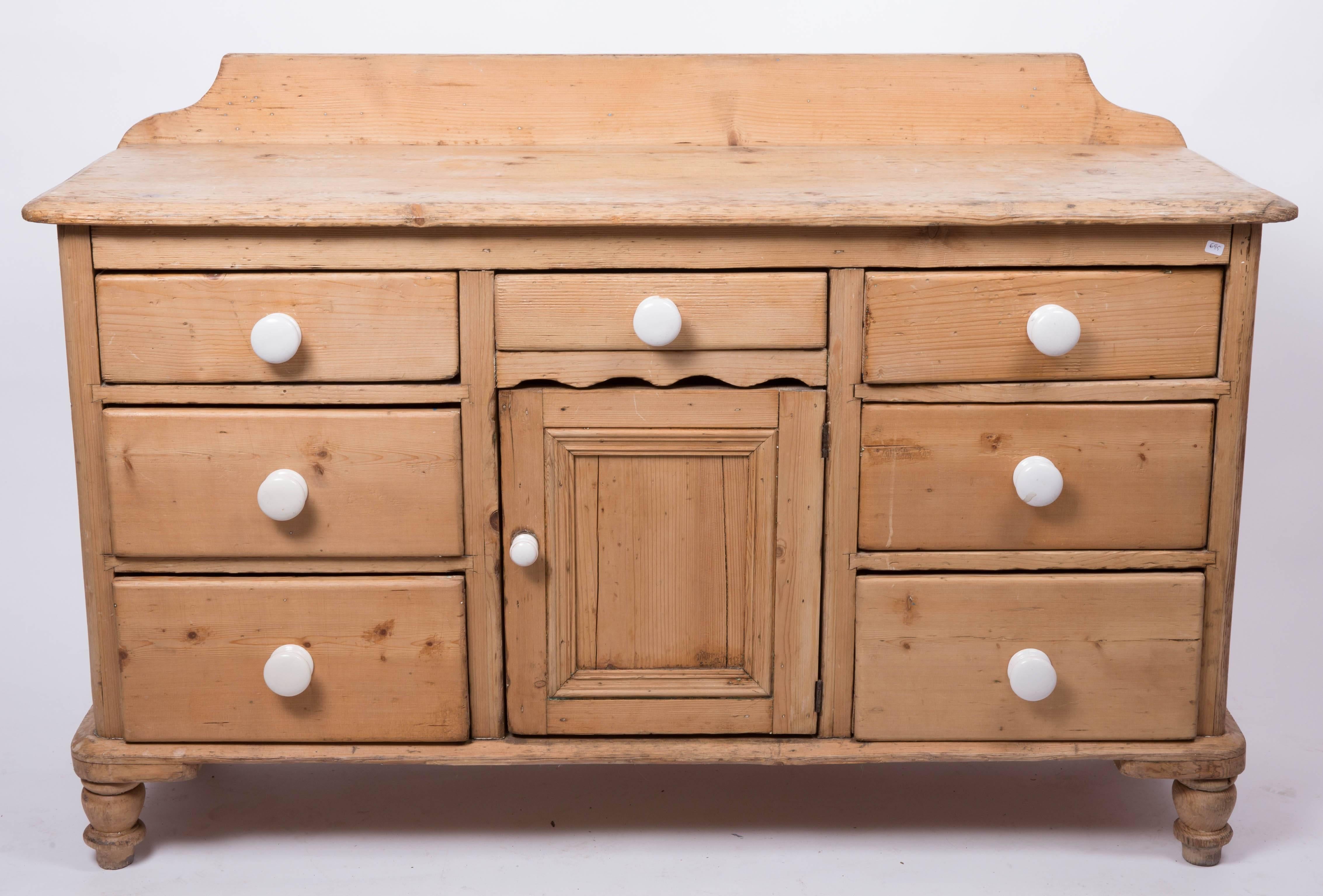 An English Country pine cupboard, console server or sideboard featuring seven drawers, each with white knobs, surrounding a recessed cupboard door and set upon ball feet.