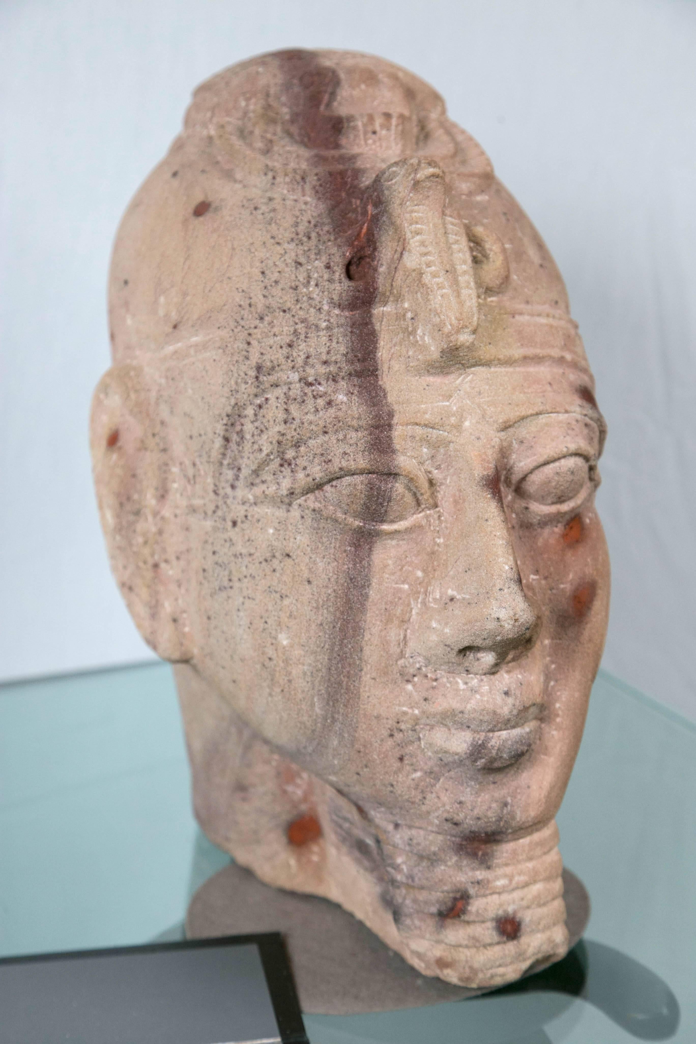 This is a reproduction, in sandstone, of an ancient head of a pharoah. He wears the cobra headress. Large eyes and full lips with the typical styled beard on his chin.
It is set on a base, attached.
    