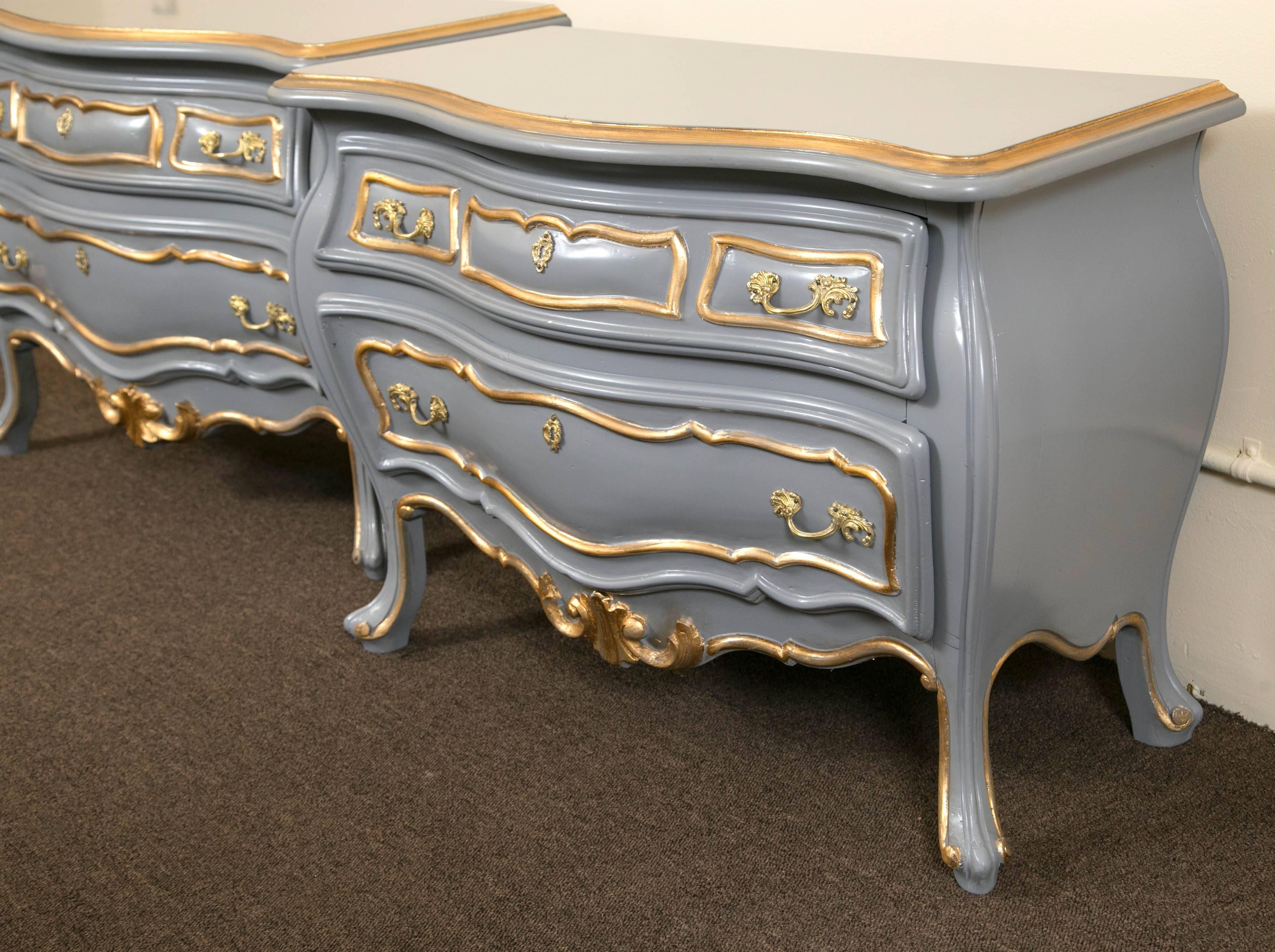 A fine Hollywood Regency pair of parcel paint and gilt decorated bombe commodes. These Louis XV style commodes or nightstands, each in a off gray color with gilt gold highlights. Bombe in form with two drawers having bronze pulls and key holes. The