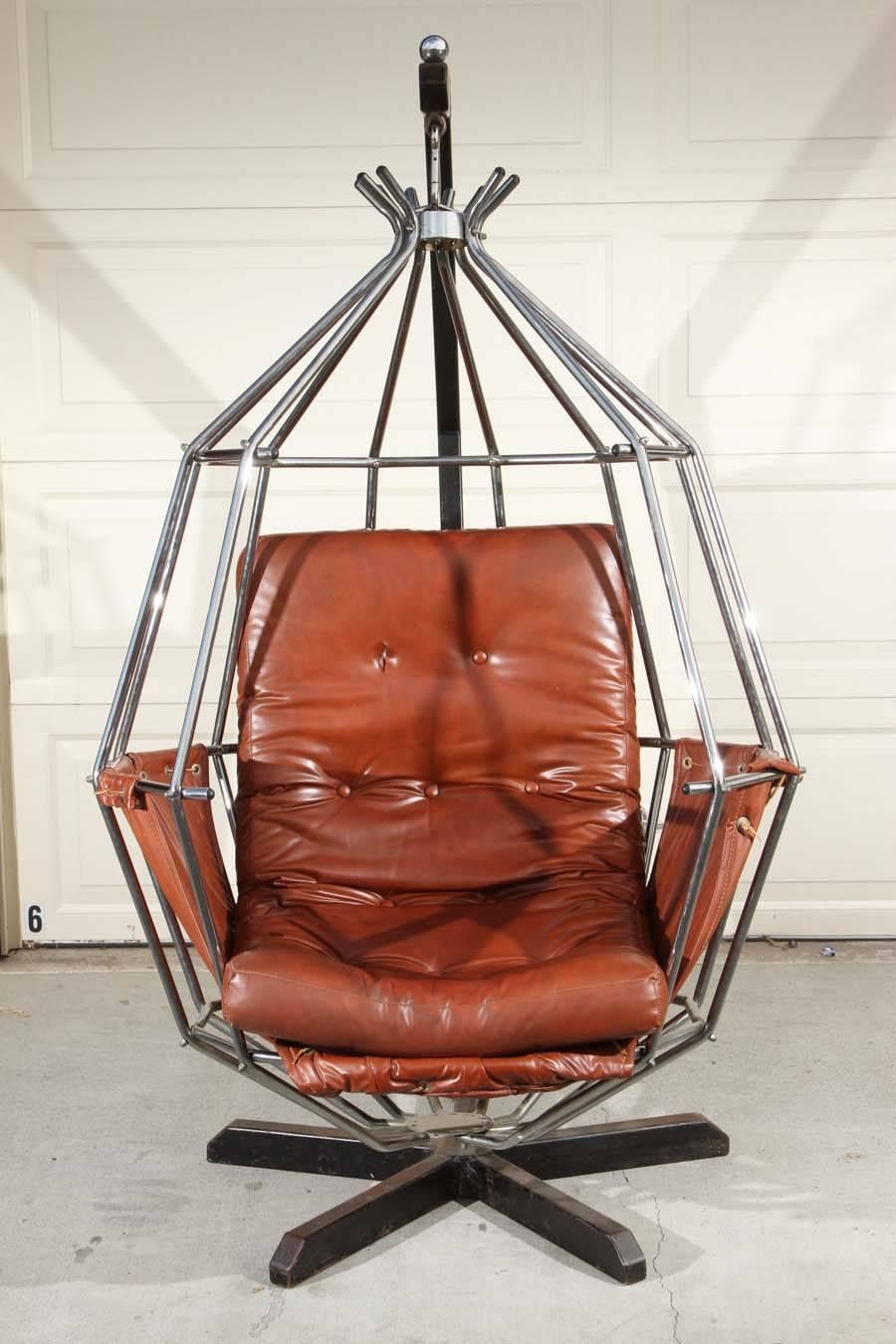 Designed by Swedish designer Ib Arberg, this iconic hanging birdcage, also known as a parrot chair was designed in 1970. The angular stand holds the steel hanging parrot chair, that holds a long cognac leather cushion, held in by a cognac leather