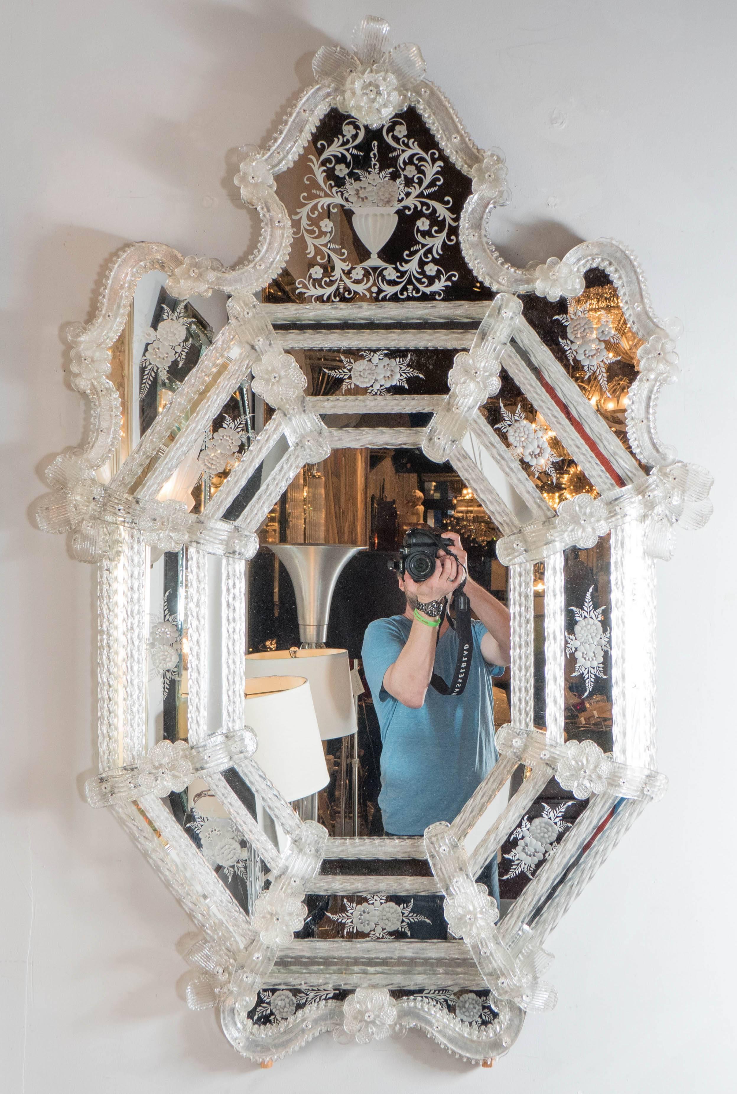 This exquisite Mid-Century Murano Venetian mirror features a crest form with scroll and reverse etched detailing throughout the borders. The mirror consists of a series of sections separated by rows of glass rods with a twist design and floral and