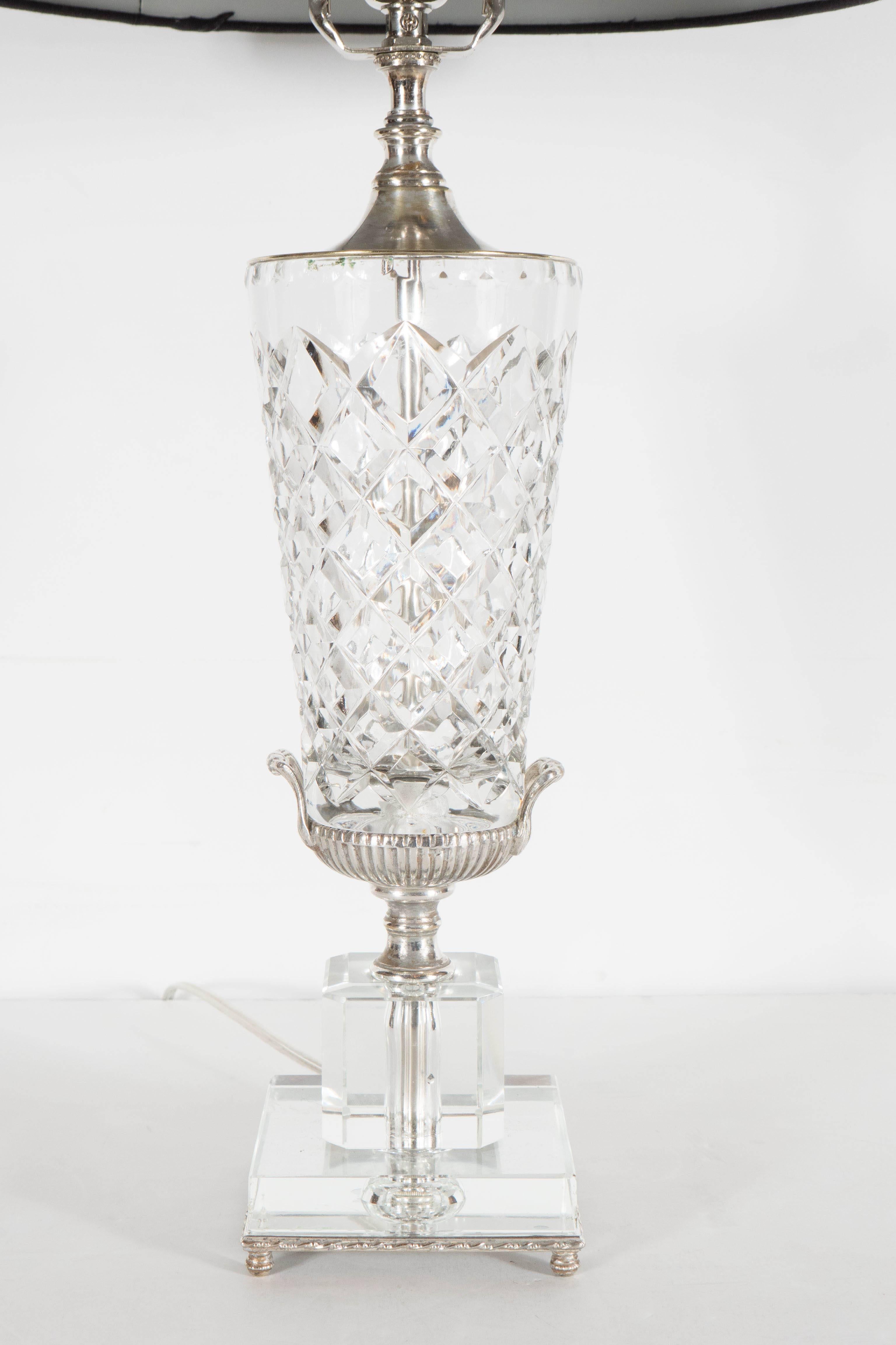 This stunning cut crystal table lamp features an urn form design. It features a silvered base with two cut crystal cube form steps supporting a silvered urn form supporting the bus crystal body of the lamp with cross hatched detailing. It has been