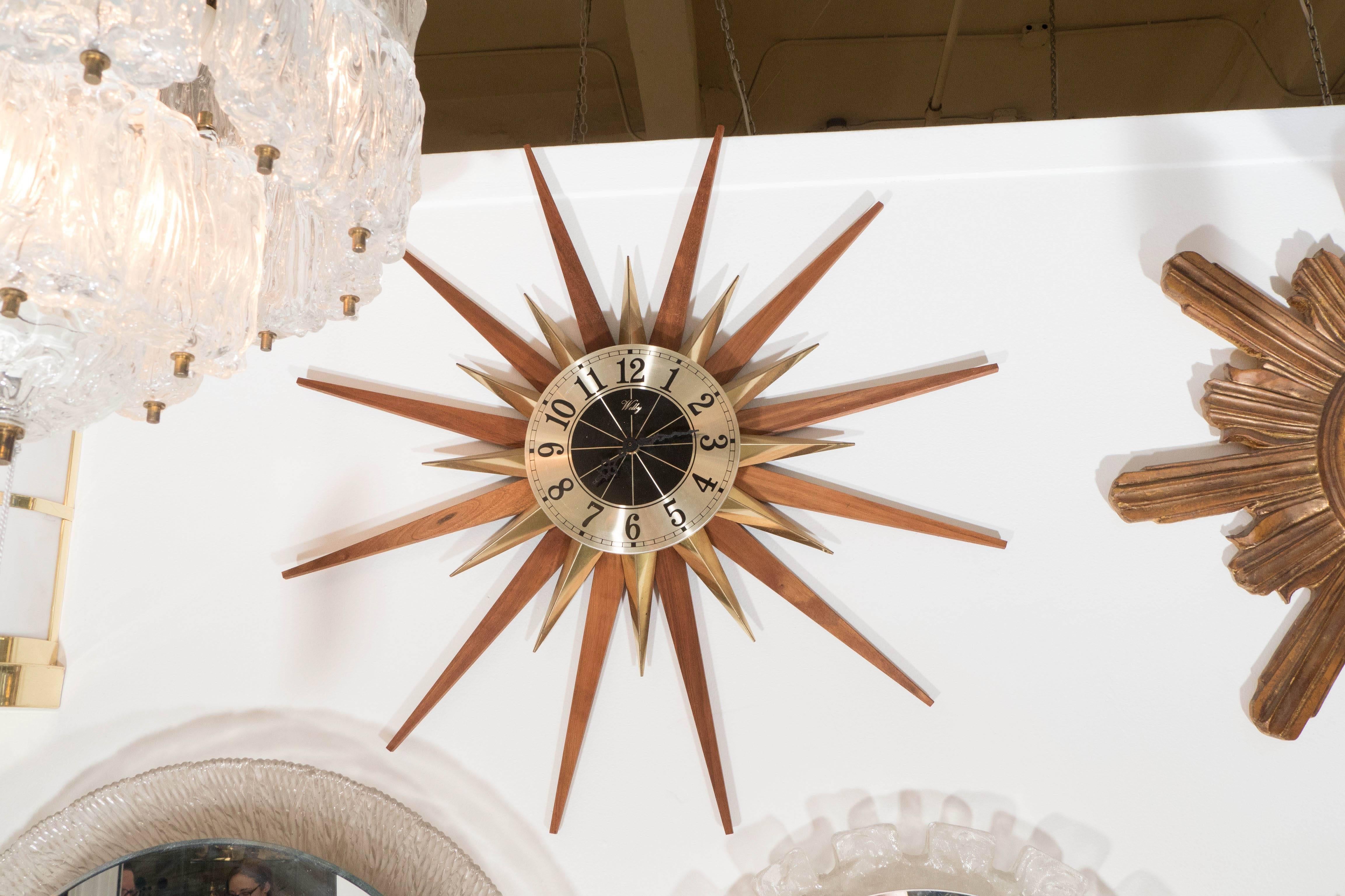 A starburst wall clock, produced by Elgin for Welby of Germany, circa 1960s-1970s, with polished brass face, surrounded by decorative rays in brass and warm toned wood. This piece is in very good vintage condition, consistent with age.