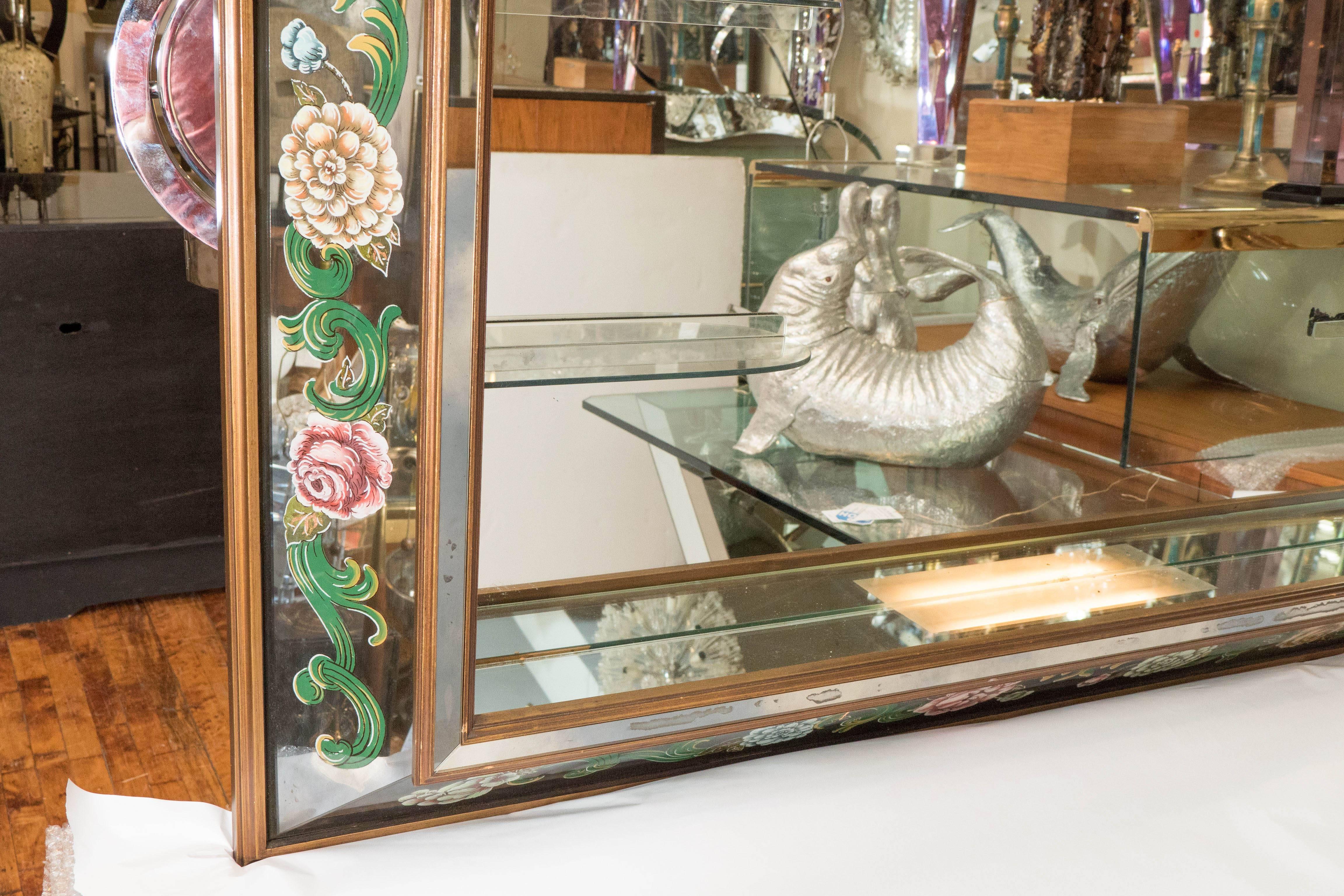 A large-scale, Hollywood Regency style wall mirror, with mirrored panel shadow box frame, bordered by reverse hand-painted flowers and acanthus leaves, inset with glass shelves. Other than presence of age appropriate wear to the frame, including