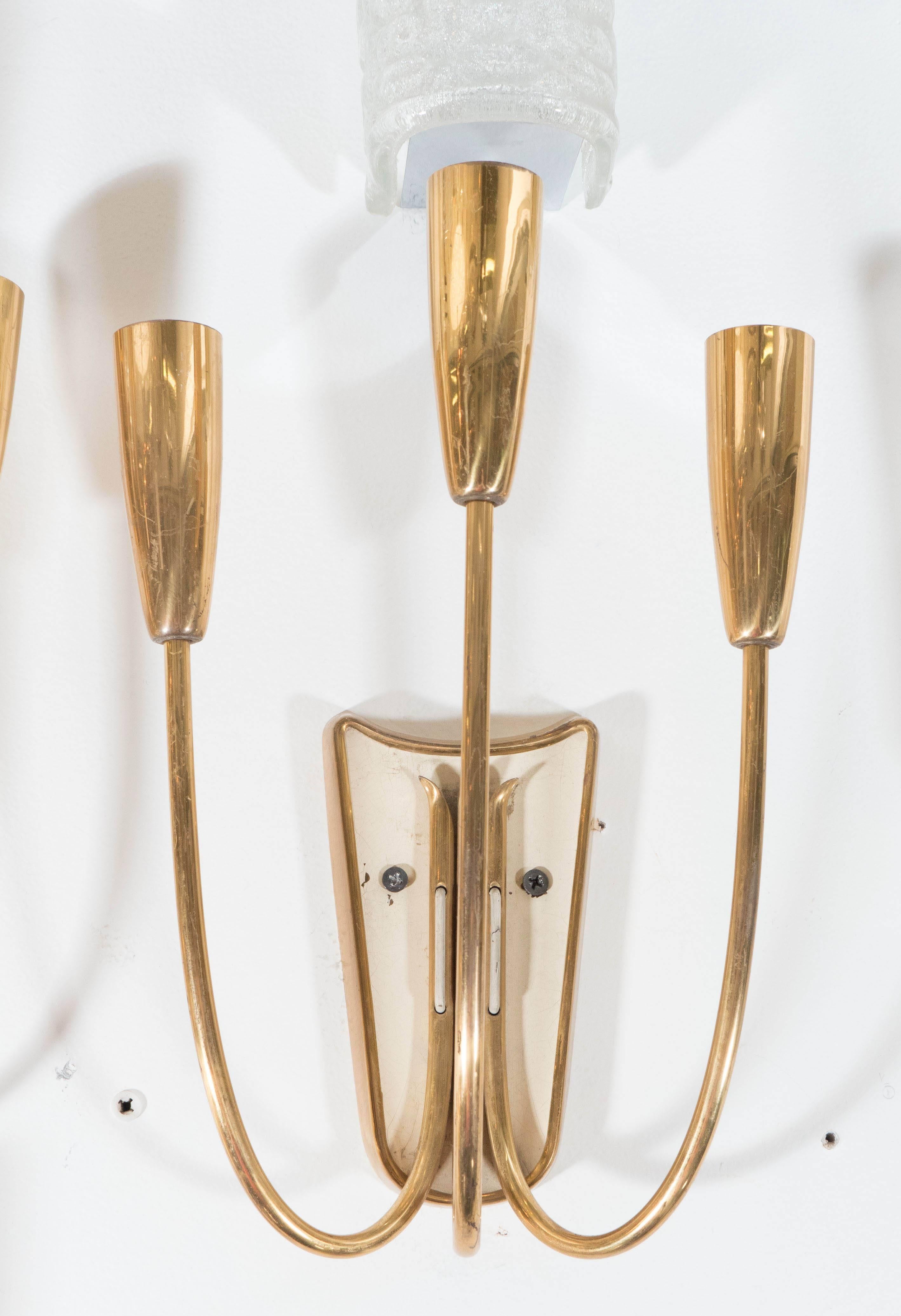 Modern style brass candelabra sconces, designed in the manner of Stilnovo, with rounded socket covers and curved stems, affixed to shield form back plates in cream colored enamel. Wiring and sockets to European standard, requires three candelabra
