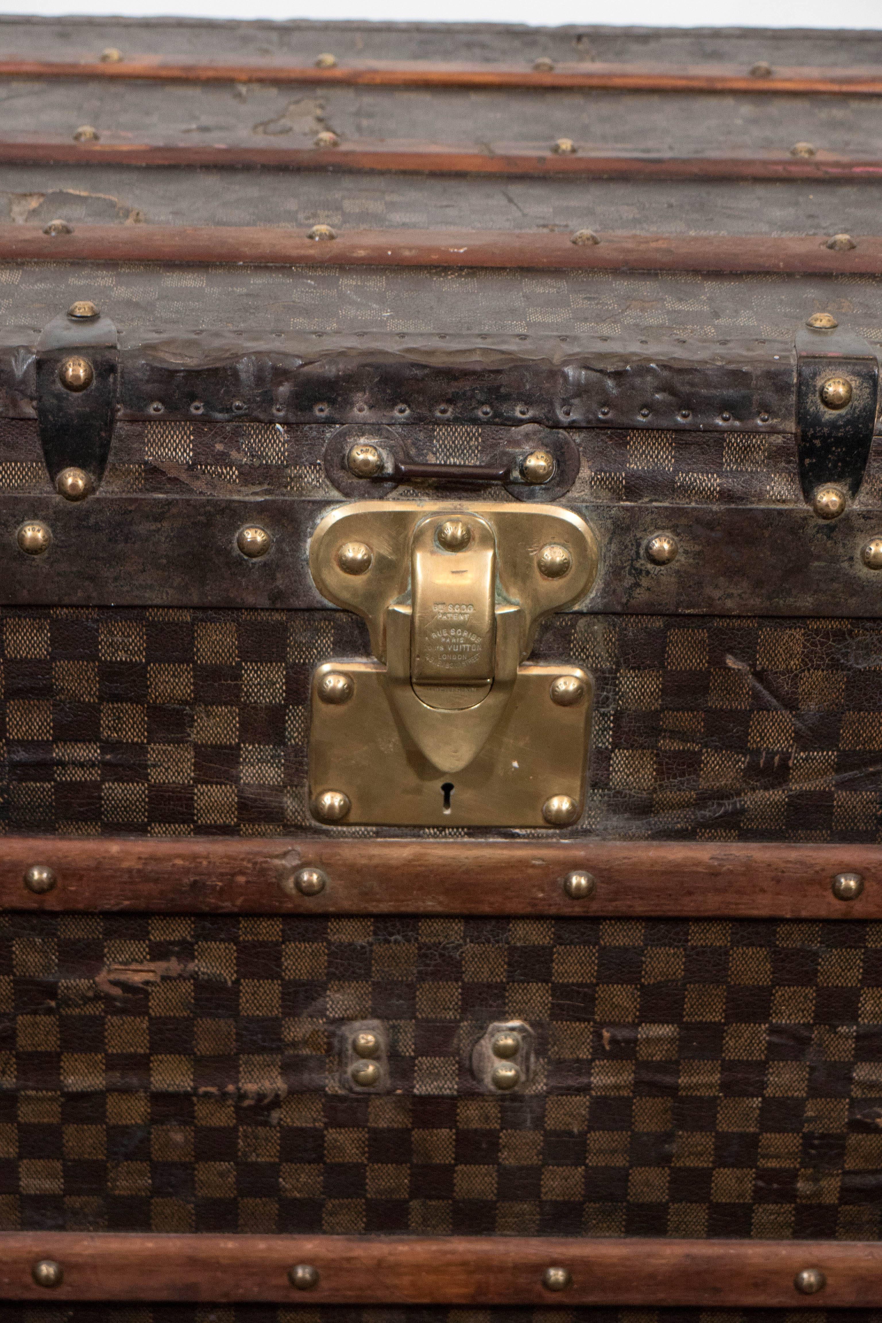 This regal French steamer trunk, produced by Louis Vuitton, circa 1889, comes in Damier pattern canvas, light brown squares to dark brown ground, flanked by iron handles, with concealed casters on the bottom; The all white fabric interior contains