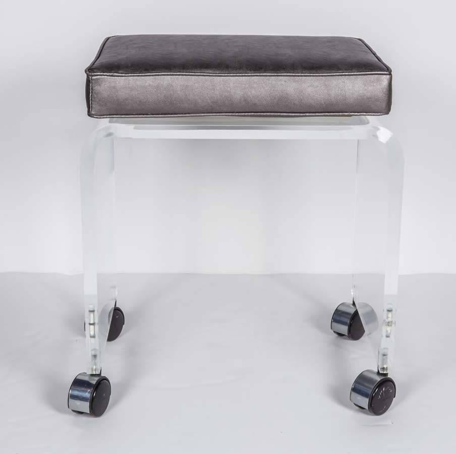 Chic and modern Lucite vanity stool with curved waterfall base design. The stool has a square seat cushion newly upholstered in metallic silver / gunmetal vegan leather. Features self welted trim along the cushion and fitted with four chromed