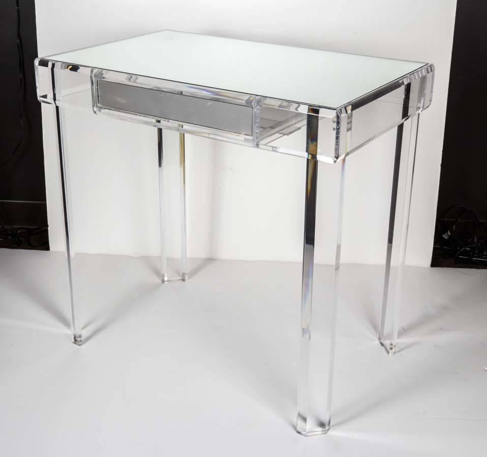 Trés chic vanity table or writing desk comprised of quality Lucite. Four Lucite column legs feature angular prism cuts, providing the illusion of cut crystal. Table surface is a single mirror and center Lucite drawer matches with front mirror and