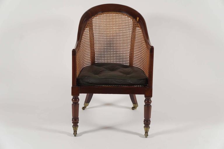 An elegant English Regency period library armchair or bergere having mahogany frame with curved crest-rail joining two rear supports, continuous caned back-rest and sides joining 'D-shaped' caned seat with tufted leather squab cushion on turned and