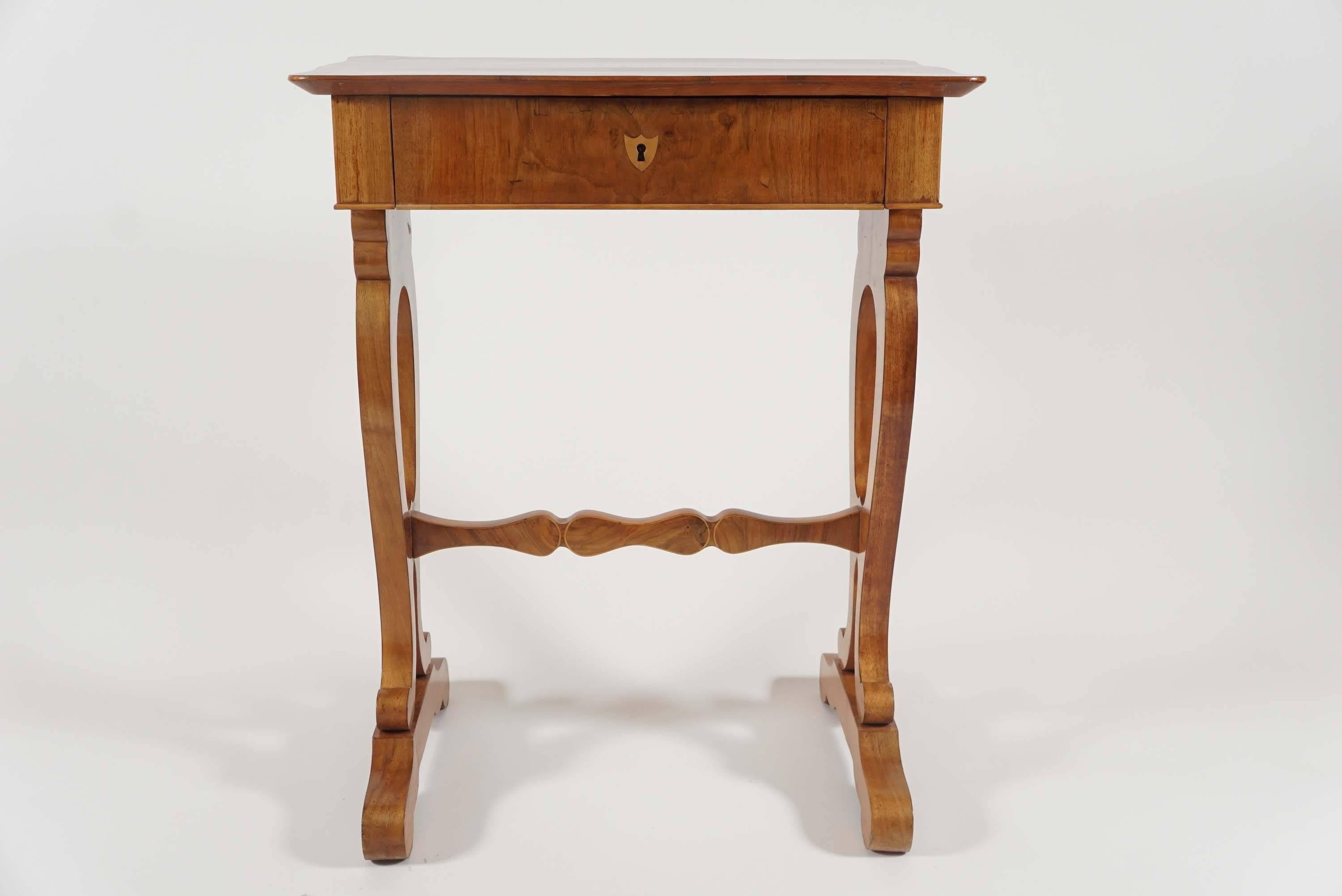 Circa 1830 Vienna, Austria, inlaid walnut Biedermeier stand, occasional, or side table having serpentine edge bookmatched flame-grained top surmounting single drawer with fitted interior and shield-inlaid key escutcheon above trestle-base with