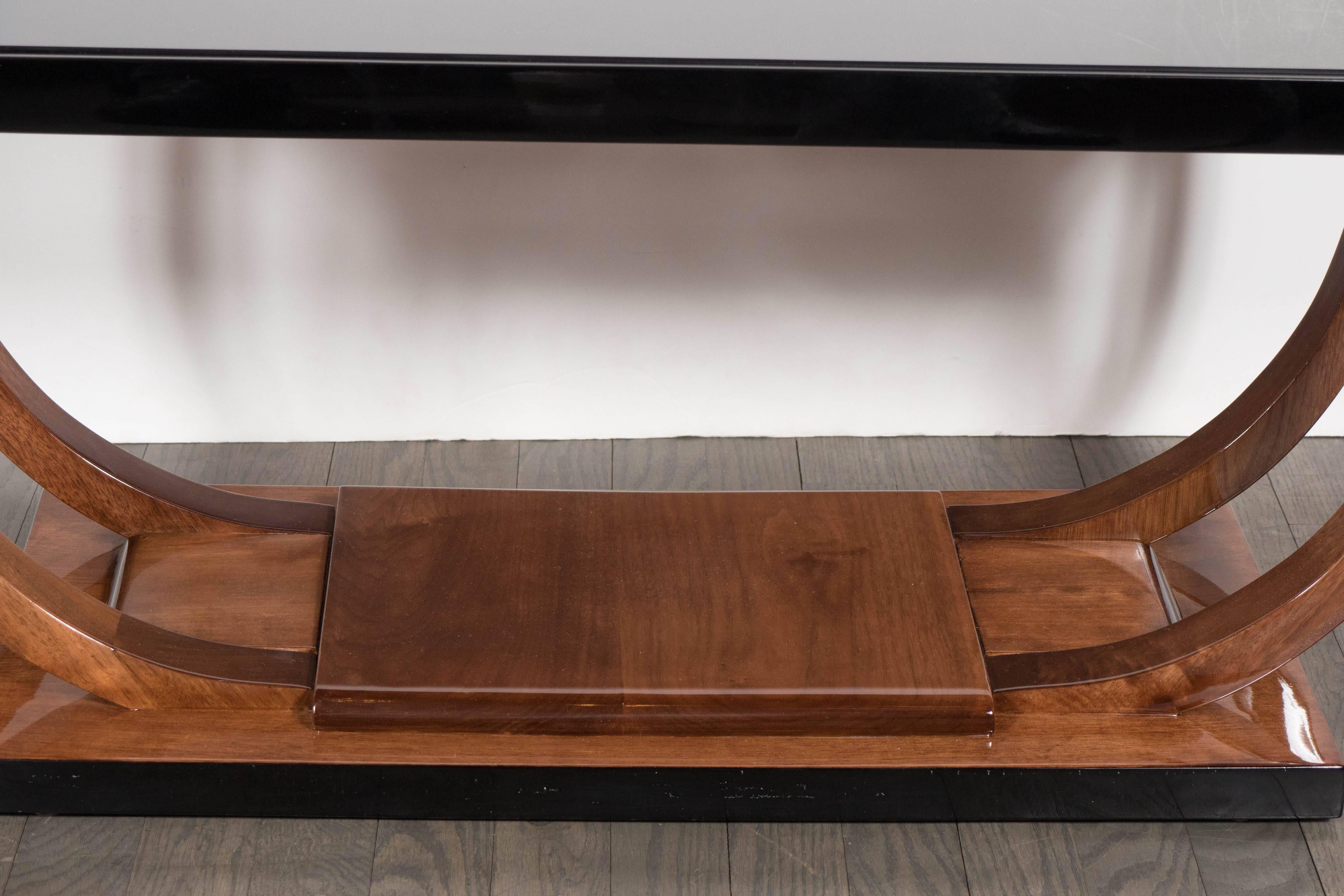 A Machine Age Art Deco cocktail table by Modernage Furniture Company featuring a banded U-form base in walnut stretching the full length of the table anchored by a rectangular base. The tabletop is trimmed in black lacquer with inset vitrolite. It