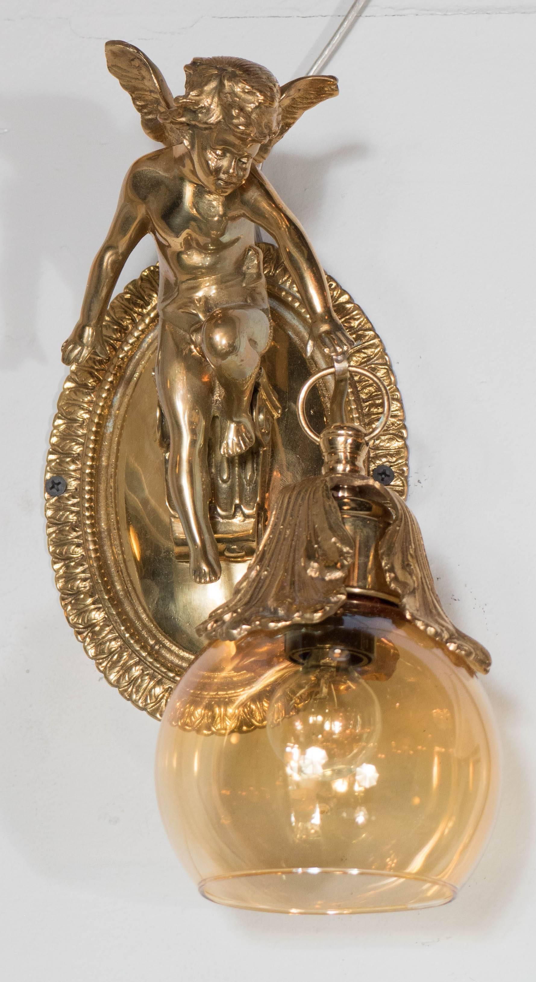 A Classic pair of brass sconces depicting winged cherubs holding a cognac hued lantern style globe draped with vine leaves. The cherub is mounted to an oval back plate bordered in a repeating laurel motif. The sconces have been newly rewired to