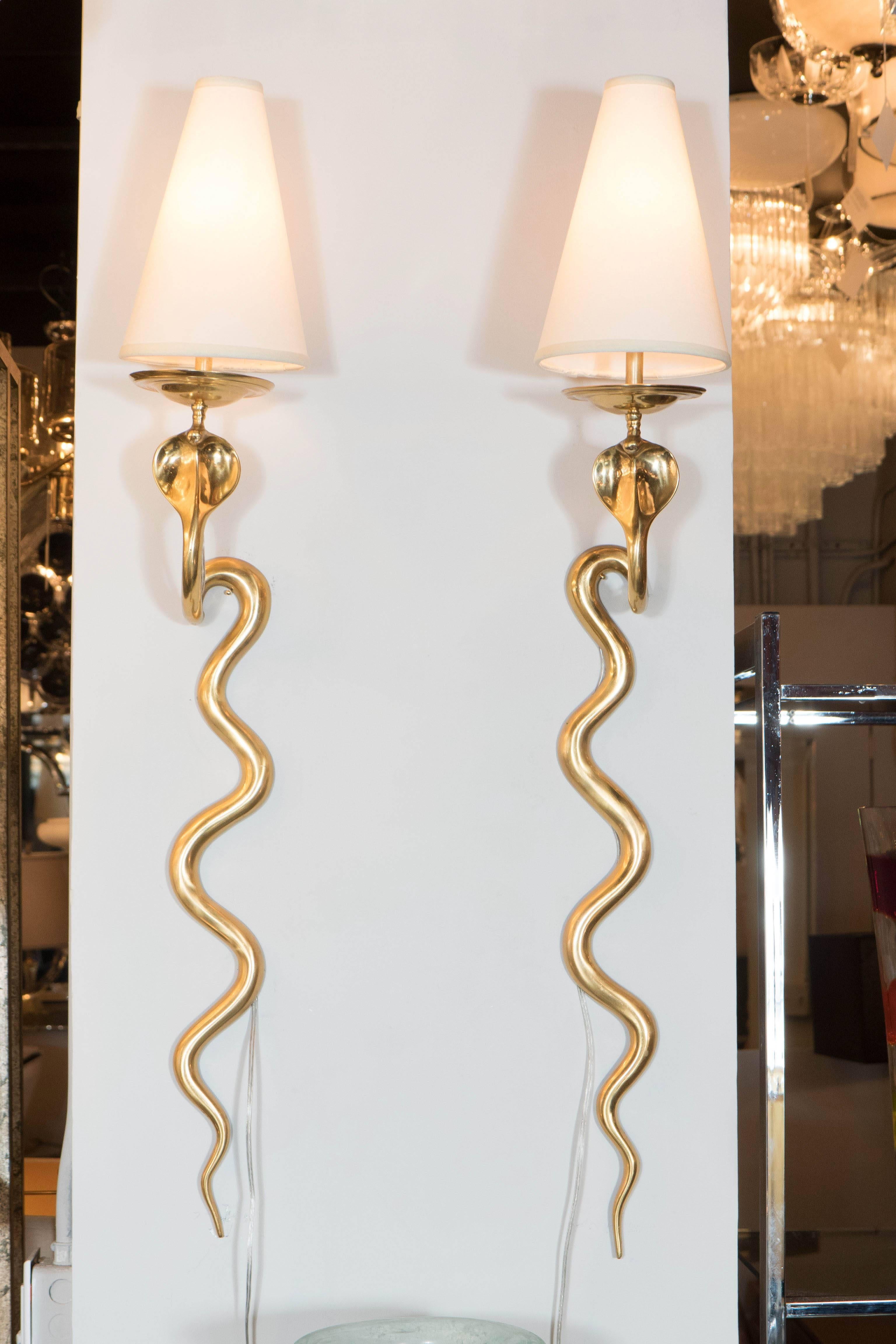 An exotic and rare pair of polished brass cobra sconces with custom shades. The snakes' tails rest on the wall while the heads, fitted with brass tongues, support candelabra sockets and conical-shaped custom white shades. An imposing pair for any