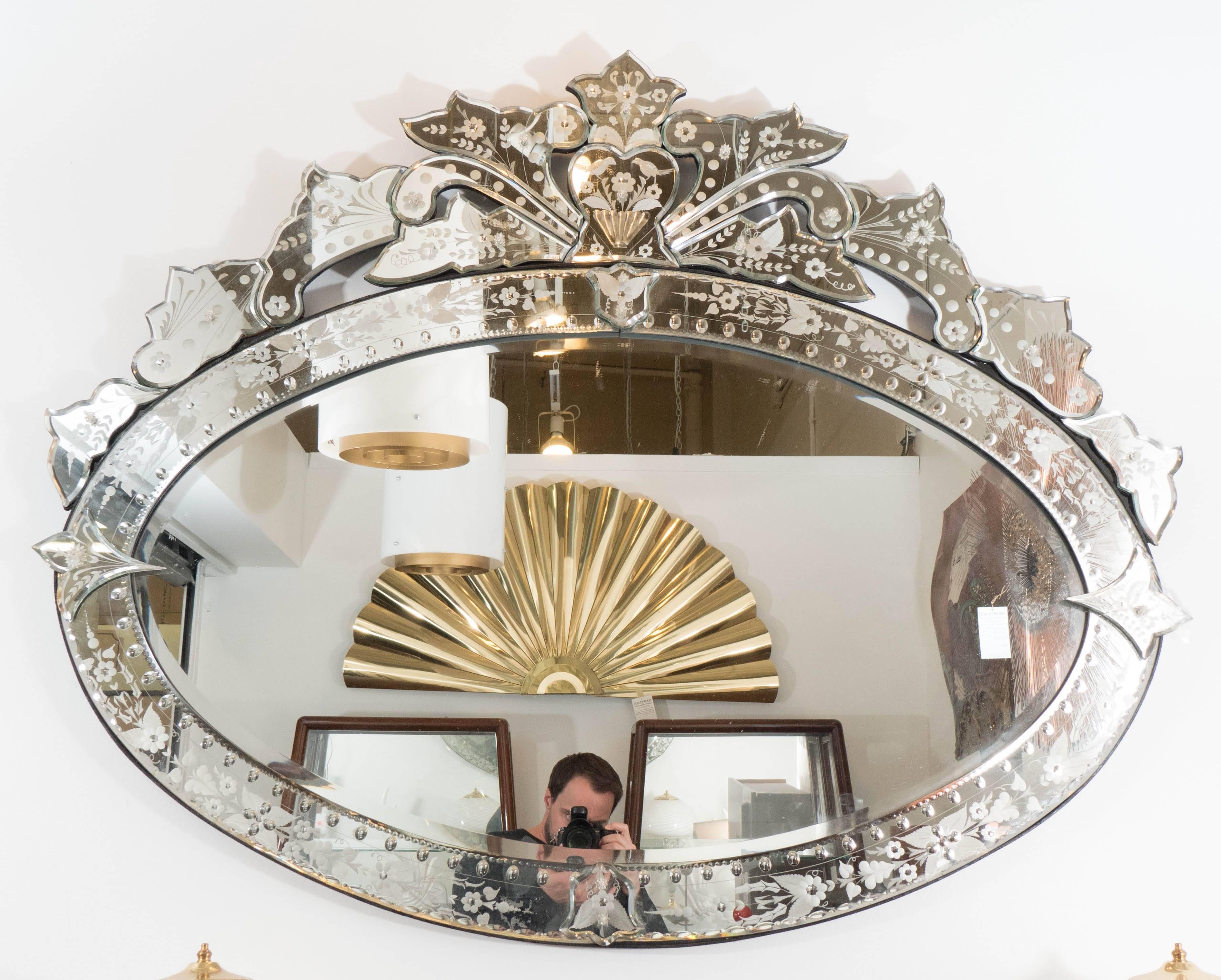 A large-scale, oval form Venetian wall mirror, produced circa 1960s, with elaborate beveled glass panel frame, detailed with convex ovals and frosted floral designs, surmounted by heart shape and surrounding acanthus, affixed by rosettes to an