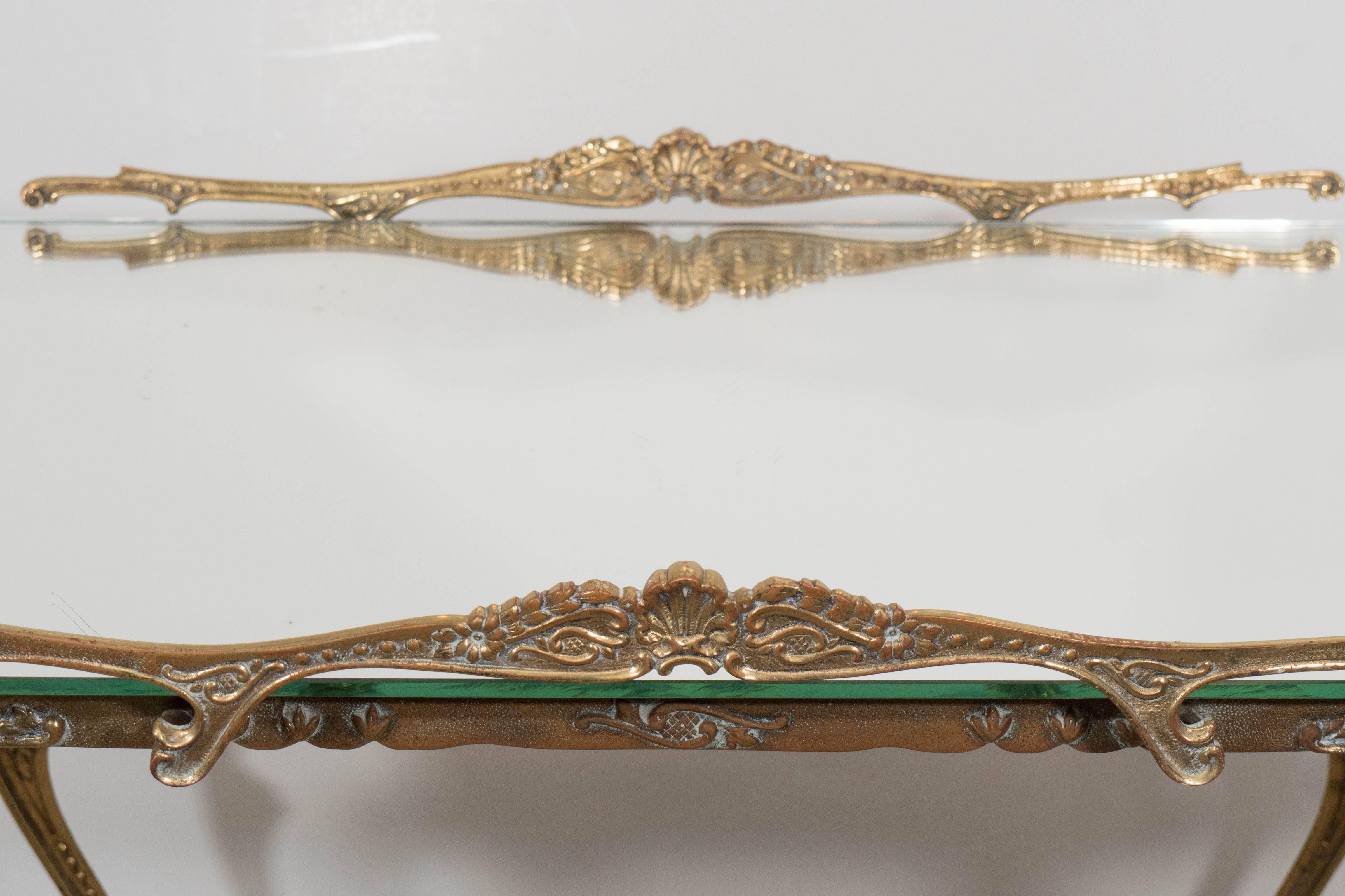 A glamorous bombe shaped, two-tier tea cart and trolley, produced circa 1950s, designed in the Louis XV style, including mirrored top, brass push handles and frame, detailed with ornate rocaille scroll-work, above lower glass tier on wheels. This