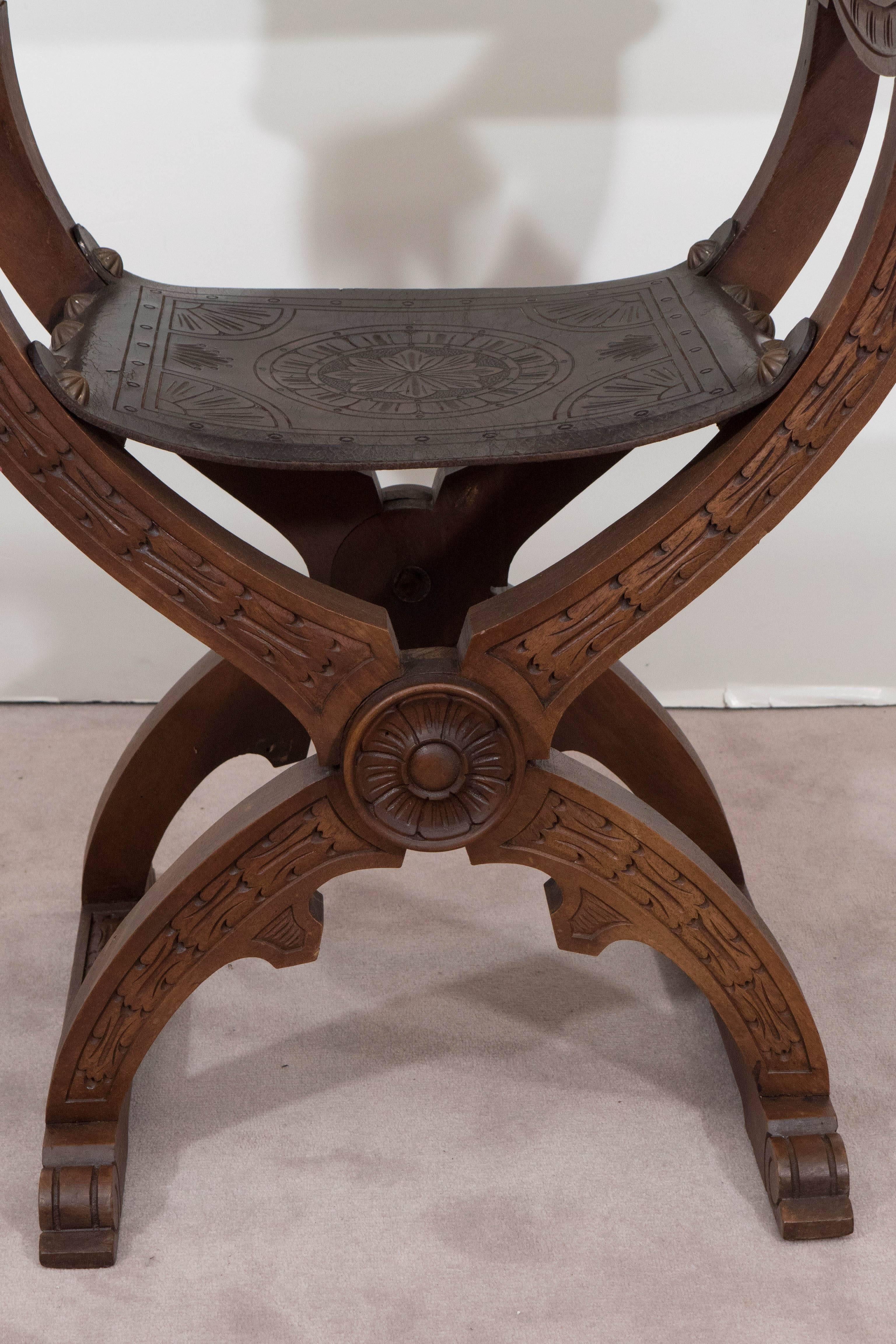 A vintage Savonarola chair, produced in Italy, circa 1950s, with beautifully detailed, etched leather seat and back, set by decorative brass nail heads against a foldable X-frame, in carved walnut with curved scroll arms. The chair remains in very