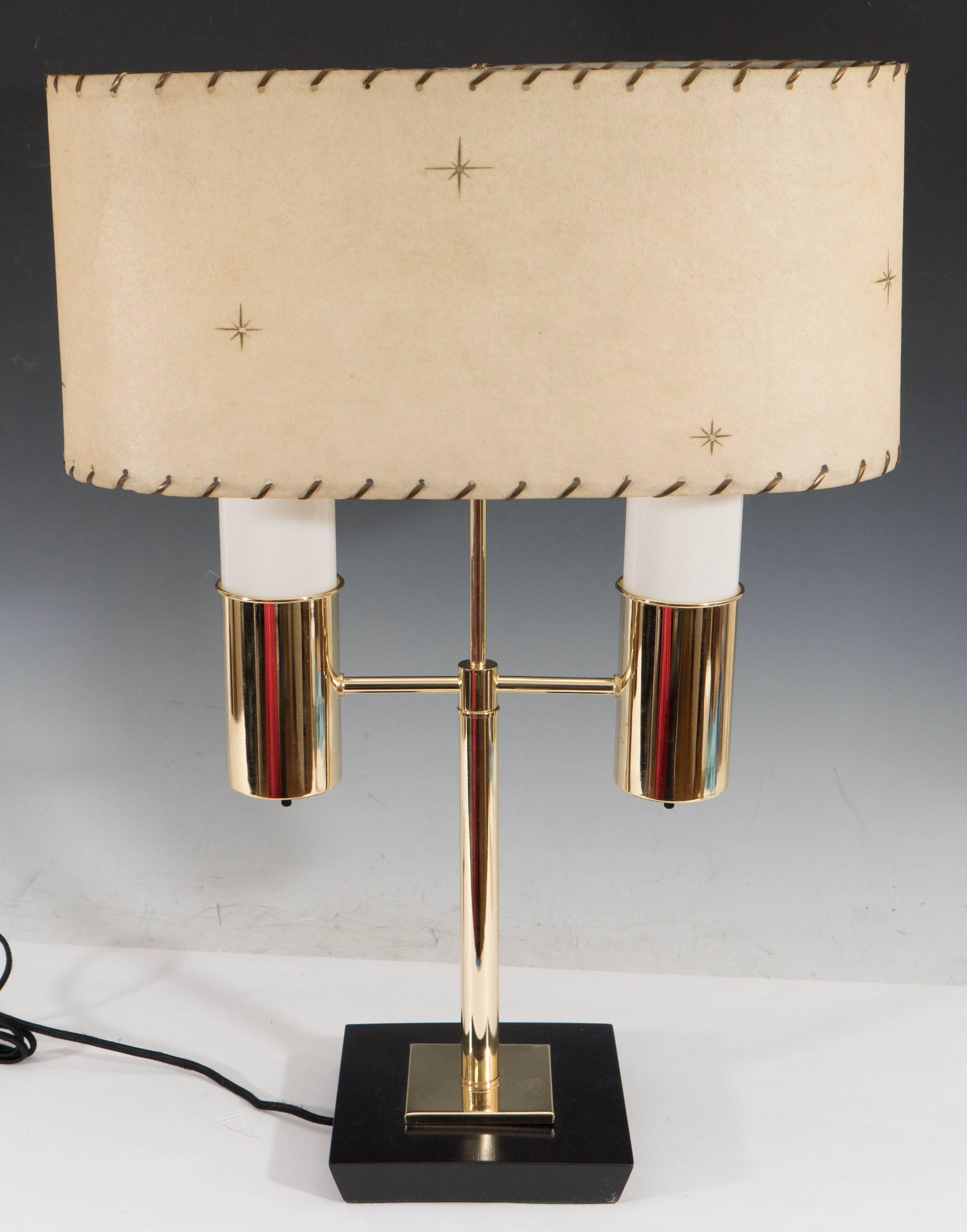 This 1950s double-light table lamp comes in polished brass, both sockets adorned with milk glass, on a black painted wood base; includes oval form parchment shade with laced stitching. The lamp remains in good vintage condition, consistent with age