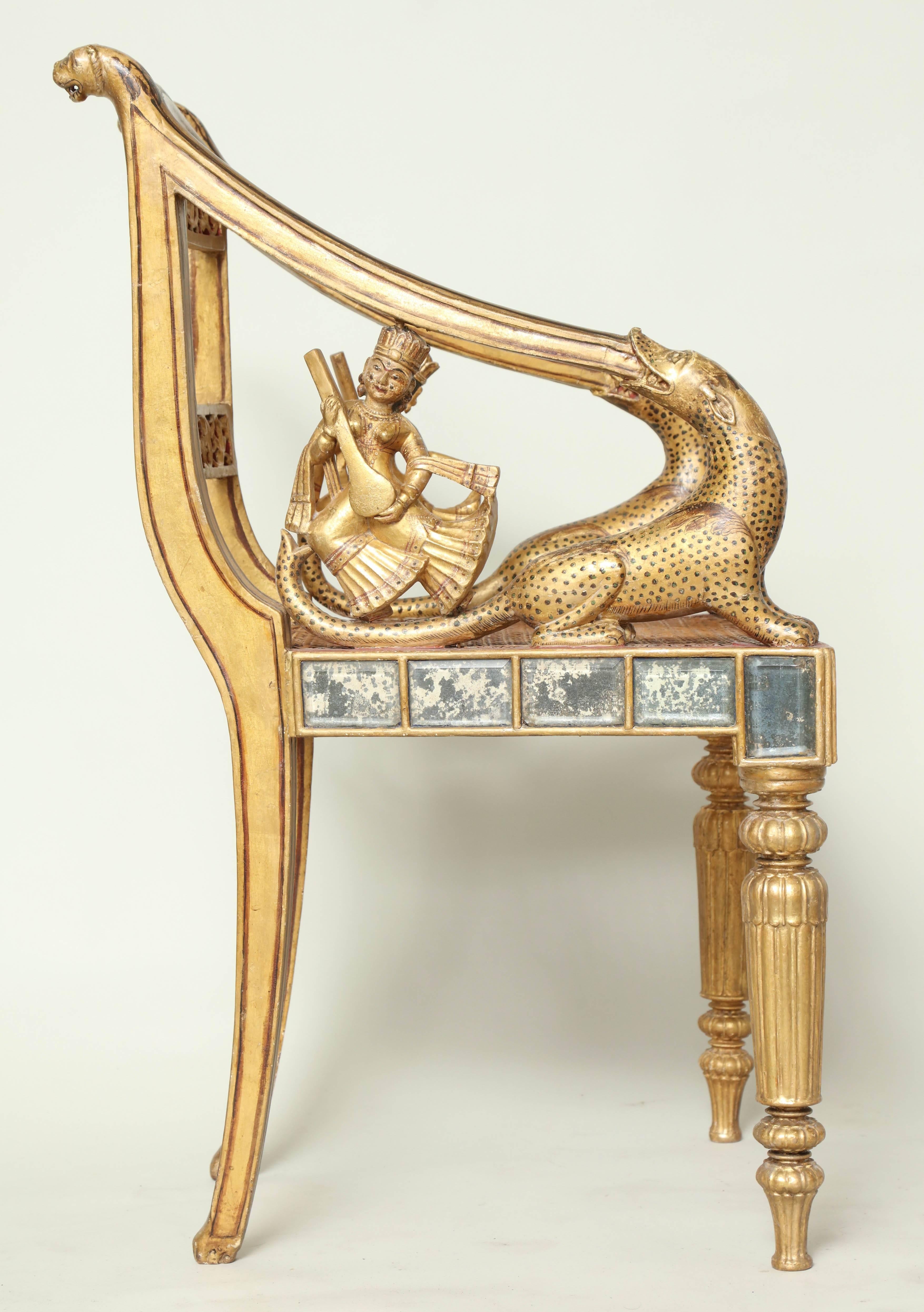 An Anglo-Siamese carved, gilt and painted armchair, having leopard carved arms with sitar playing deities, the pierced back rails with central medallion with deity and surrounding foliate decoration with leopard head tops, over inset bevelled mirror