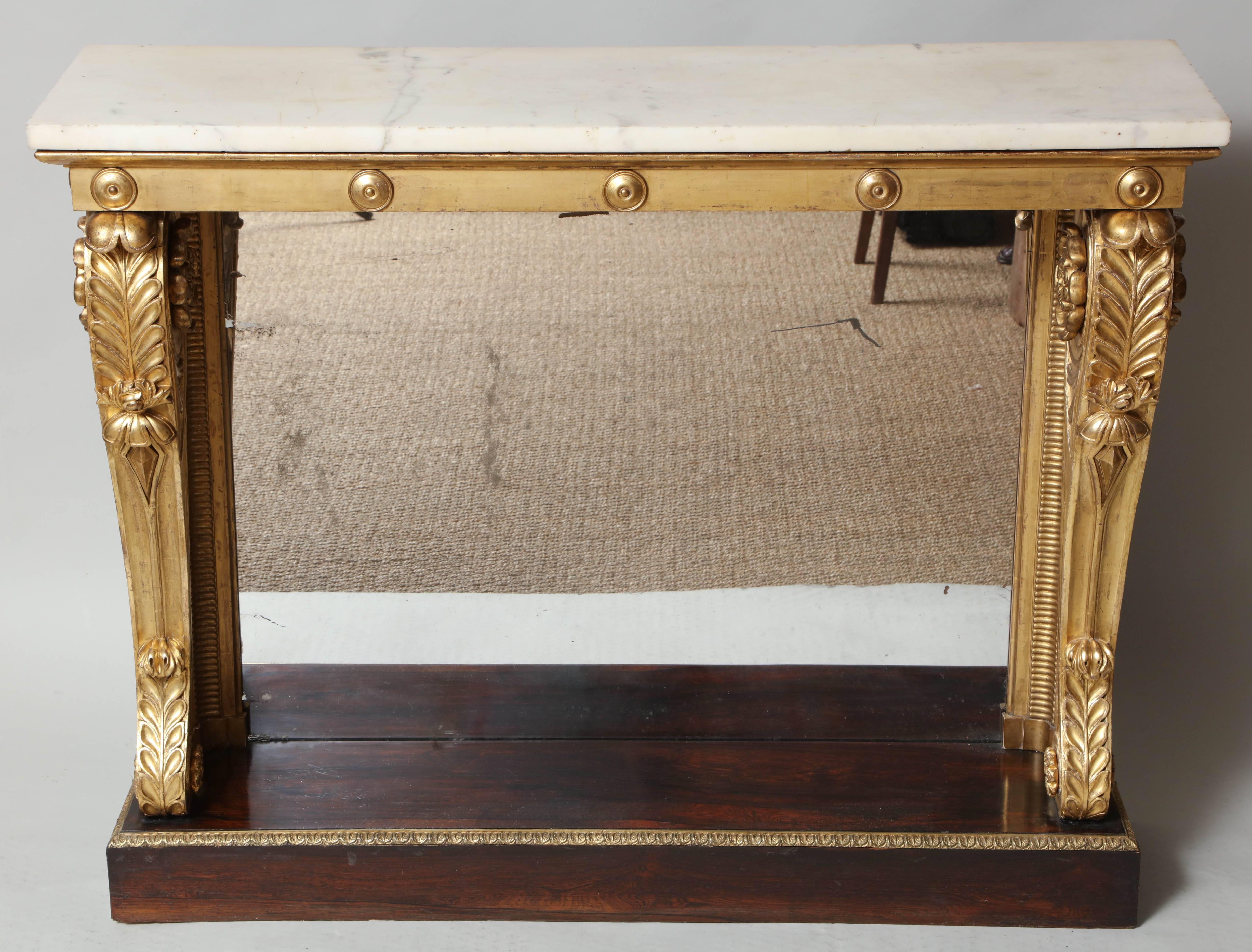 Very fine early 19th century pier table of useful narrow proportions, the original white marble top over giltwood apron with applied bosses, the bold scrolled legs with acanthus leaf and rosette motifs standing on rich rosewood plinth with gilt