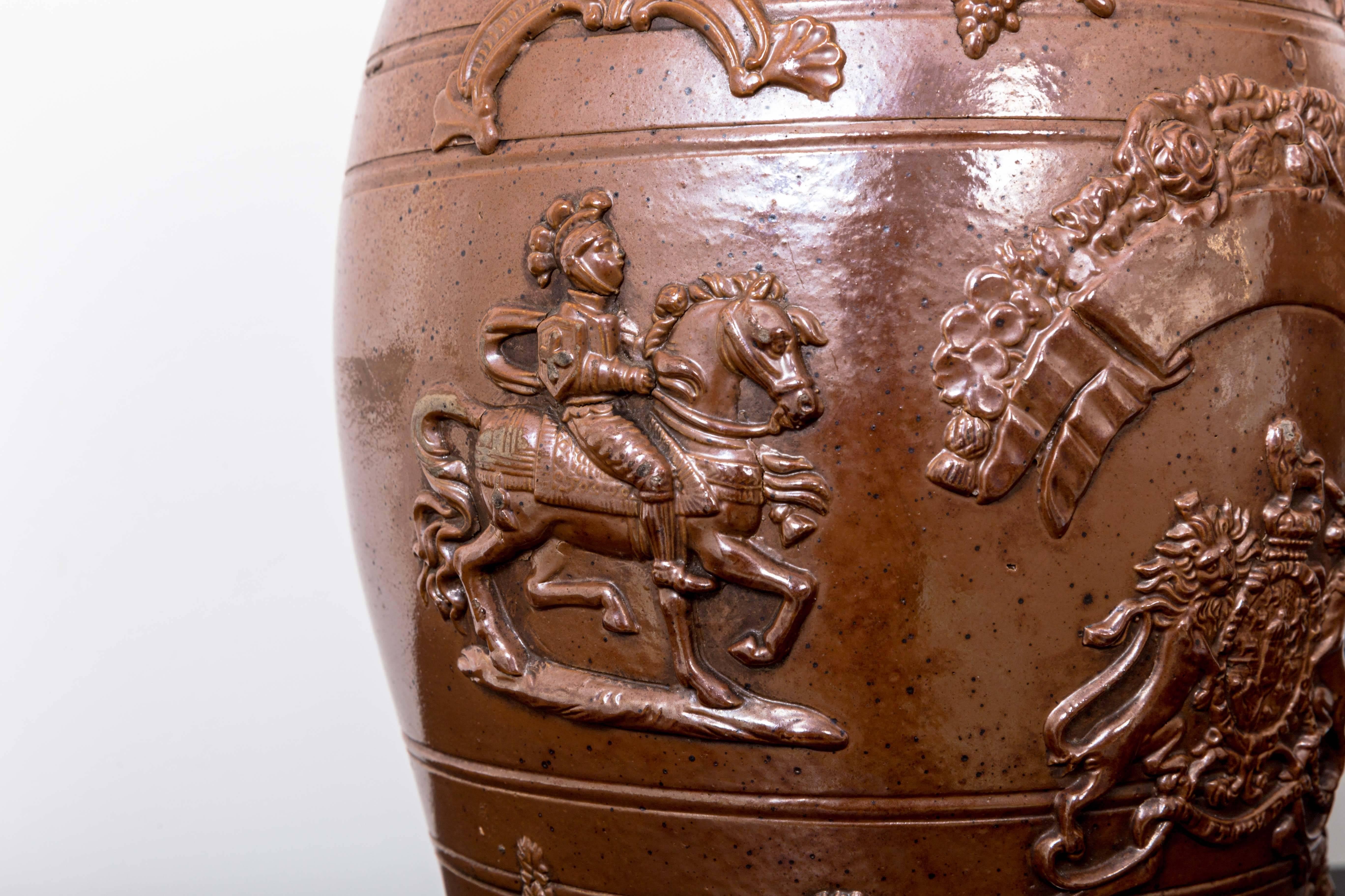 English salt glazed stoneware spirits dispenser, barrel form, decorated with bas-relief motifs; including royal coat of arms, knights on horseback, lions, banner and garland with wooden spigot and cork top.