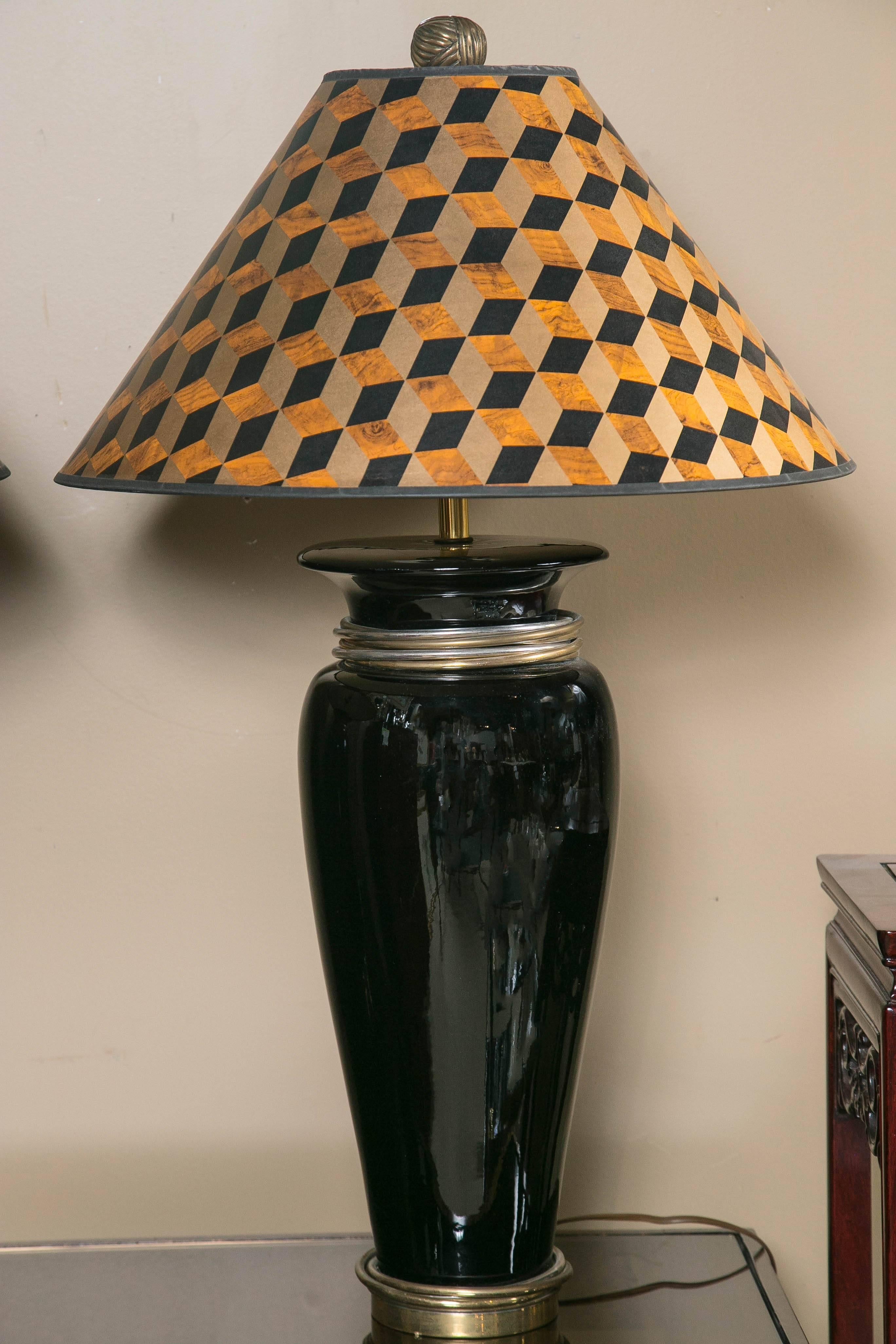 Black ceramic urn form lamps with tonal metal loose rings at neck and base, retaining their original optical grid patterned paper shades, substantial knob finials, labels and wiring, which is in good working order.