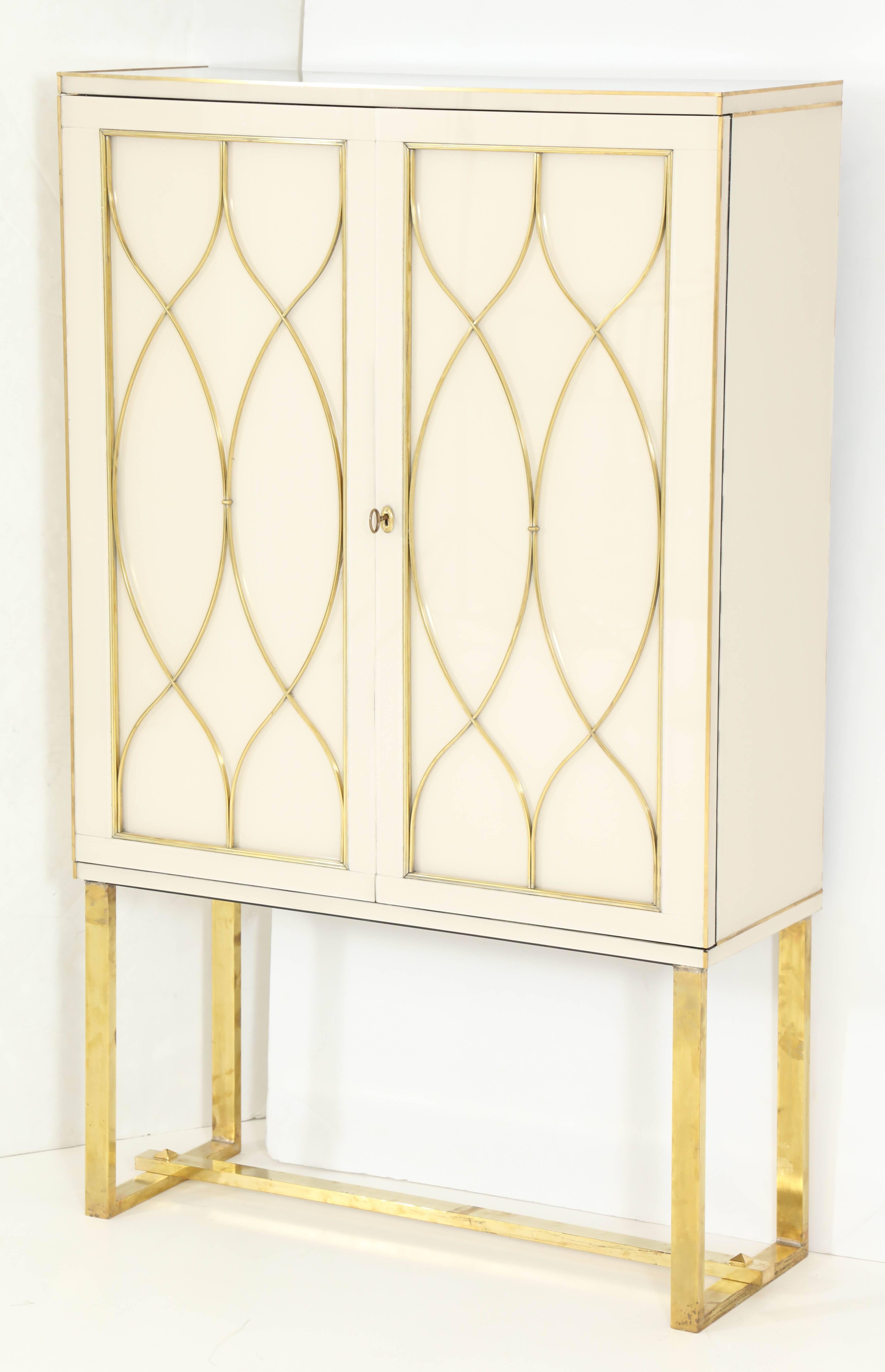 This Italian Mid-Century tall cabinet or dry bar is absolutely exquisite in its detail and craftmanship. Made of wood and covered in a high-gloss ivory colored venetian Opaline glass with beautiful brass inlays and brass flat front legs. Working