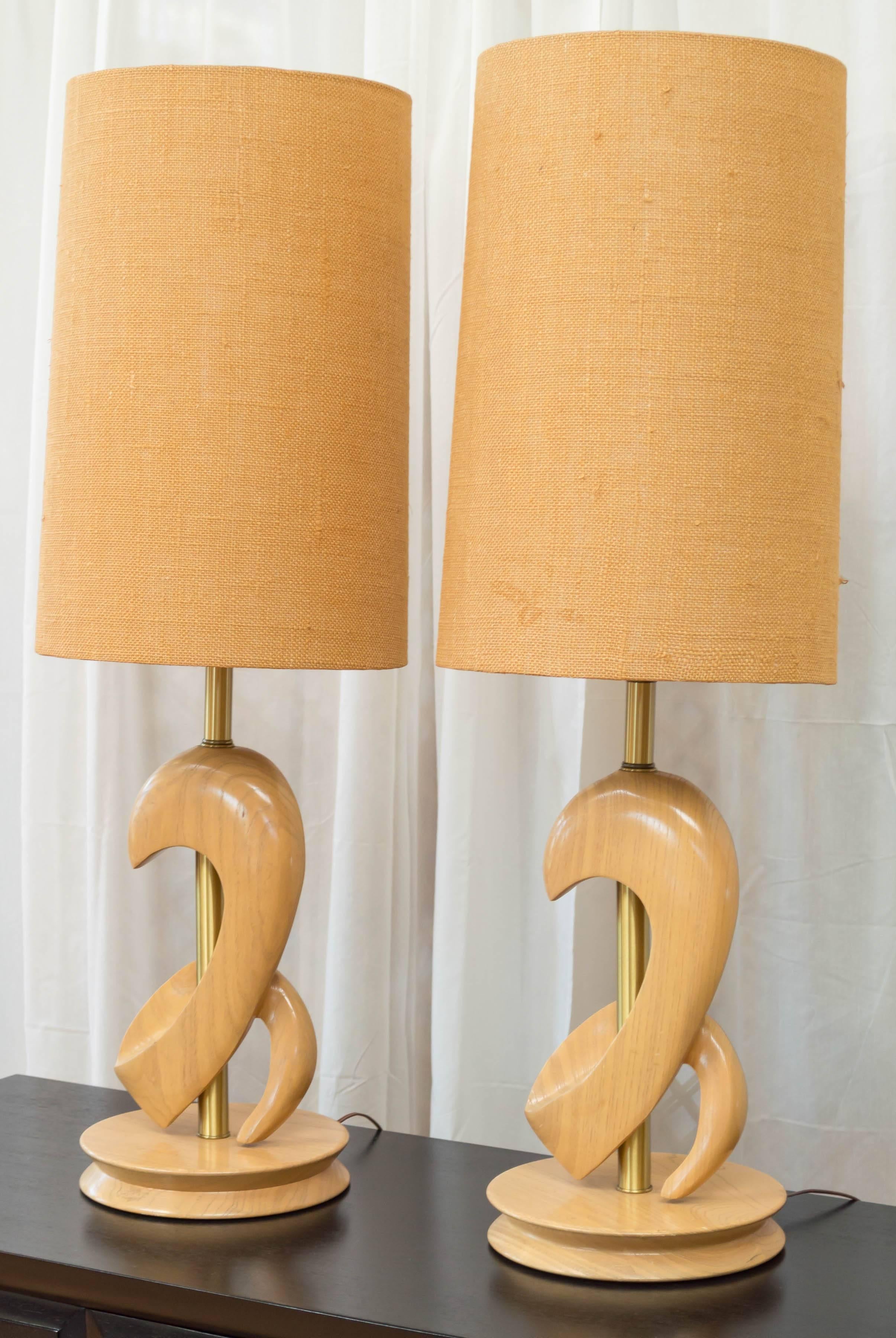 A pair of Atomic Age bleached oak table lamps with original burlap shades attributed to designer Yasha Heifetz.

Hand-carved free-form body flows banner-like around its brass stem, and poses upon a broad pulley-edge base. The lamp’s thick curves