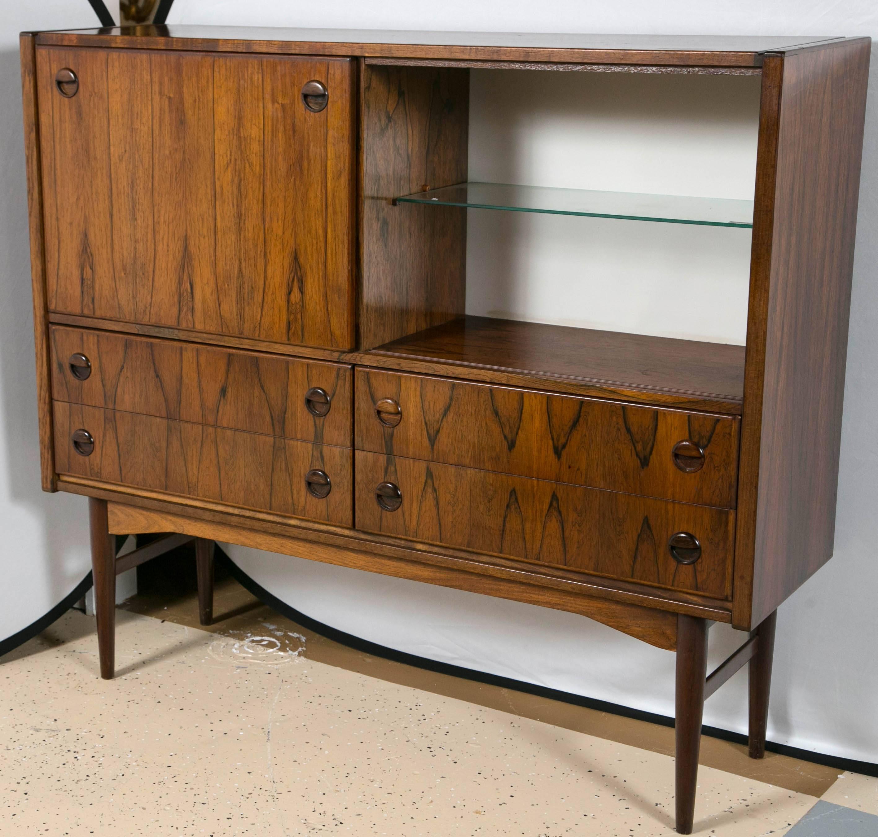 A custom quality Scandinavian Mid-Century rosewood bar cabinet. The casing sitting on tapering legs of Mid-Century Modern design. The undercarriage supporting four lower drawers and an upper rosewood interior and glass fitted area to accompany