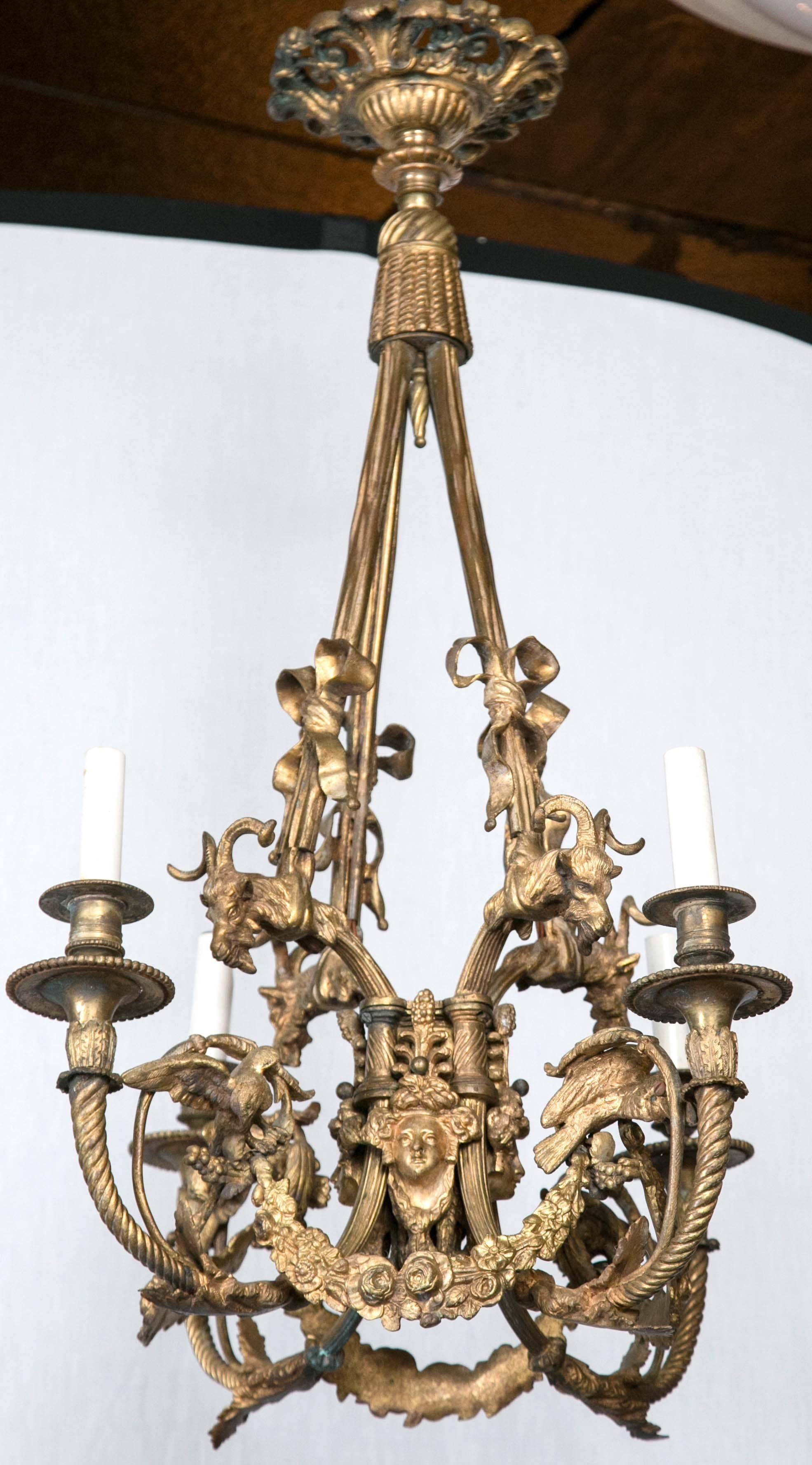 The top a medallion form ceiling mount. Above a tassel form to which are connected four suspending arms, each decorated with a bow, terminating with the top of the candle arms with goat heads. The arms swoop down and up, and are twisted. Between