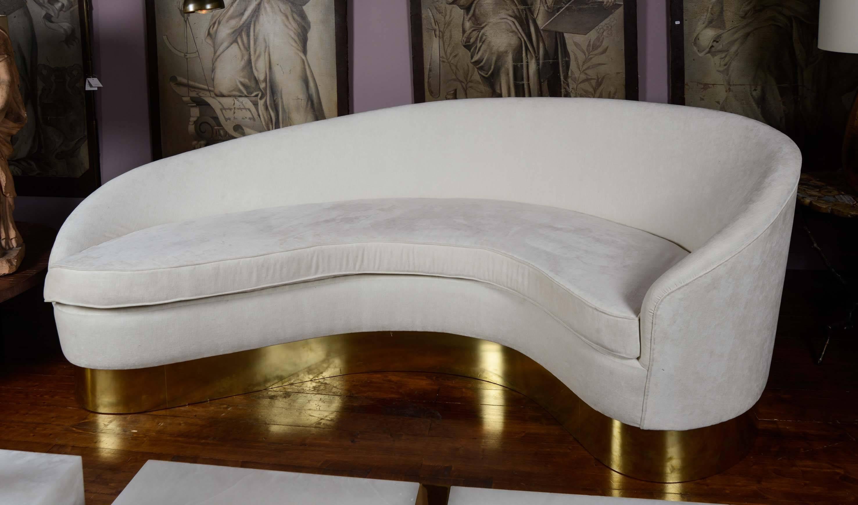 Curved sofa upholstered with ecru fabric, cushions, brass base.