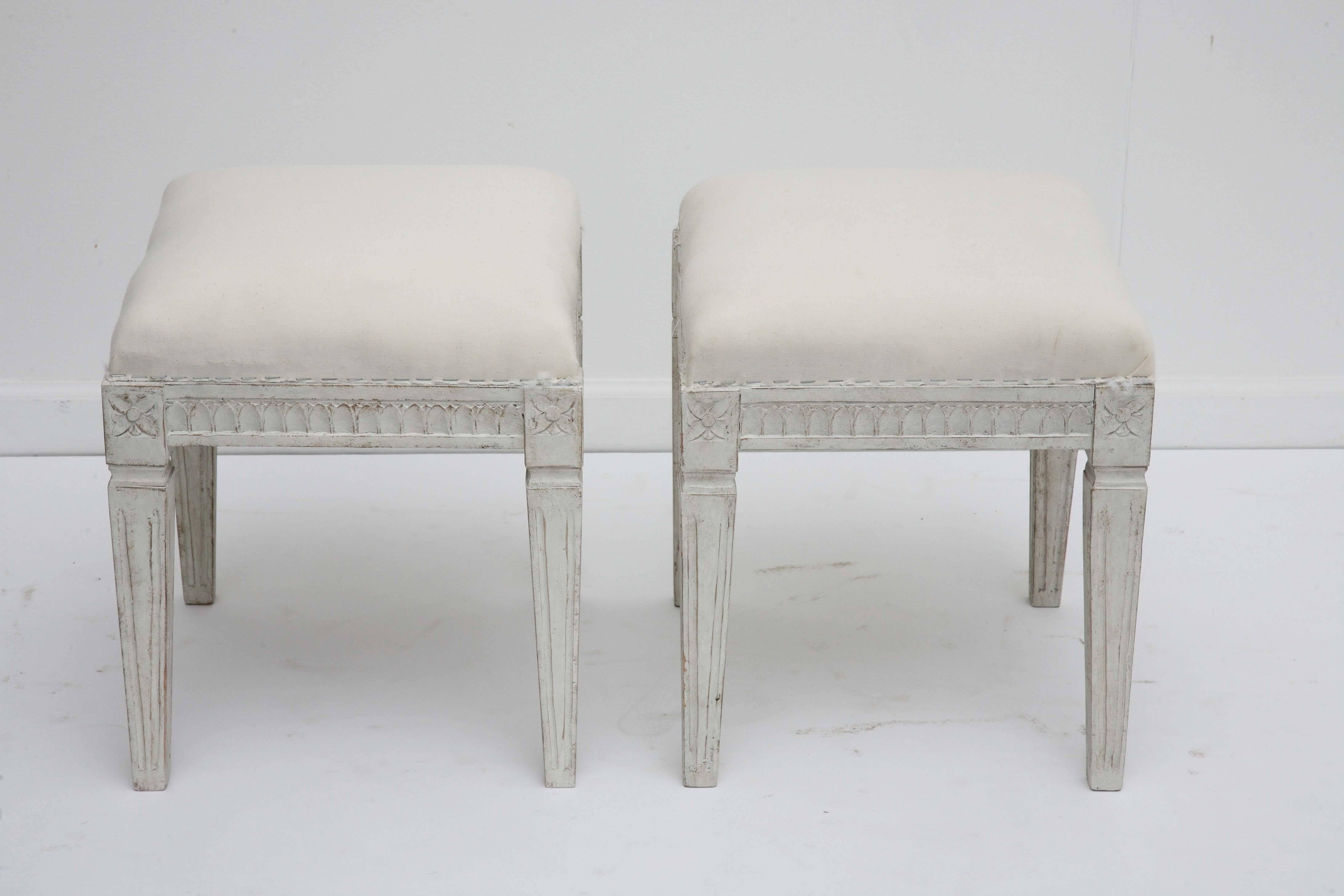 A pair of antique Swedish Gustavian painted stools, with carved lamb's tongue apron, square tapered legs with carved flower rosettes at top of each leg; they are painted in white-grayish distressed finish , curved upholstered tops.

19th