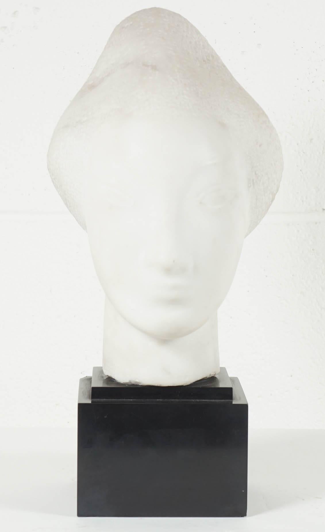 Here is a beautiful sculpture of a female head mounted on a solid black stone base. The piece is highly stylized and the formed hair has a textured quality that contrasts the smooth surface of the face. There is an incised signature on the side of