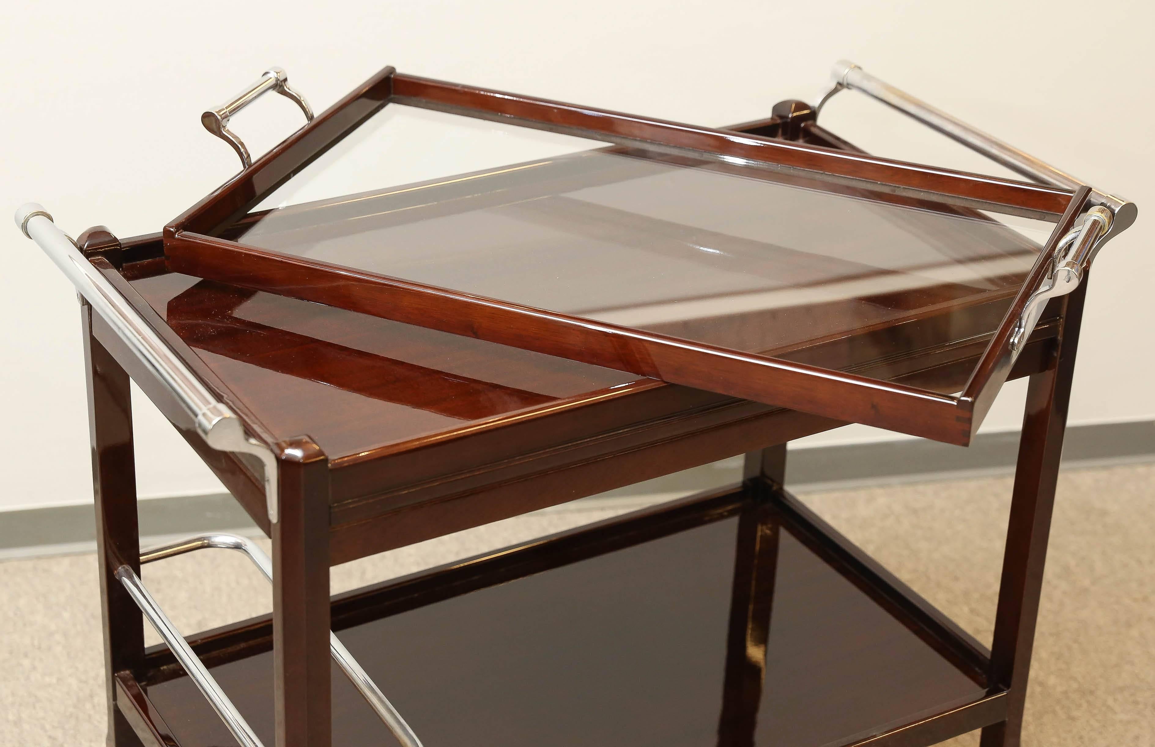 A wooden frame of the bar cart supports two wooden trays. One glass removable tray with small chrome handles is located on the top of the upper shelf. On the bottom, wooden legs are connected with chrome tubes. Four small wheels are attached to the
