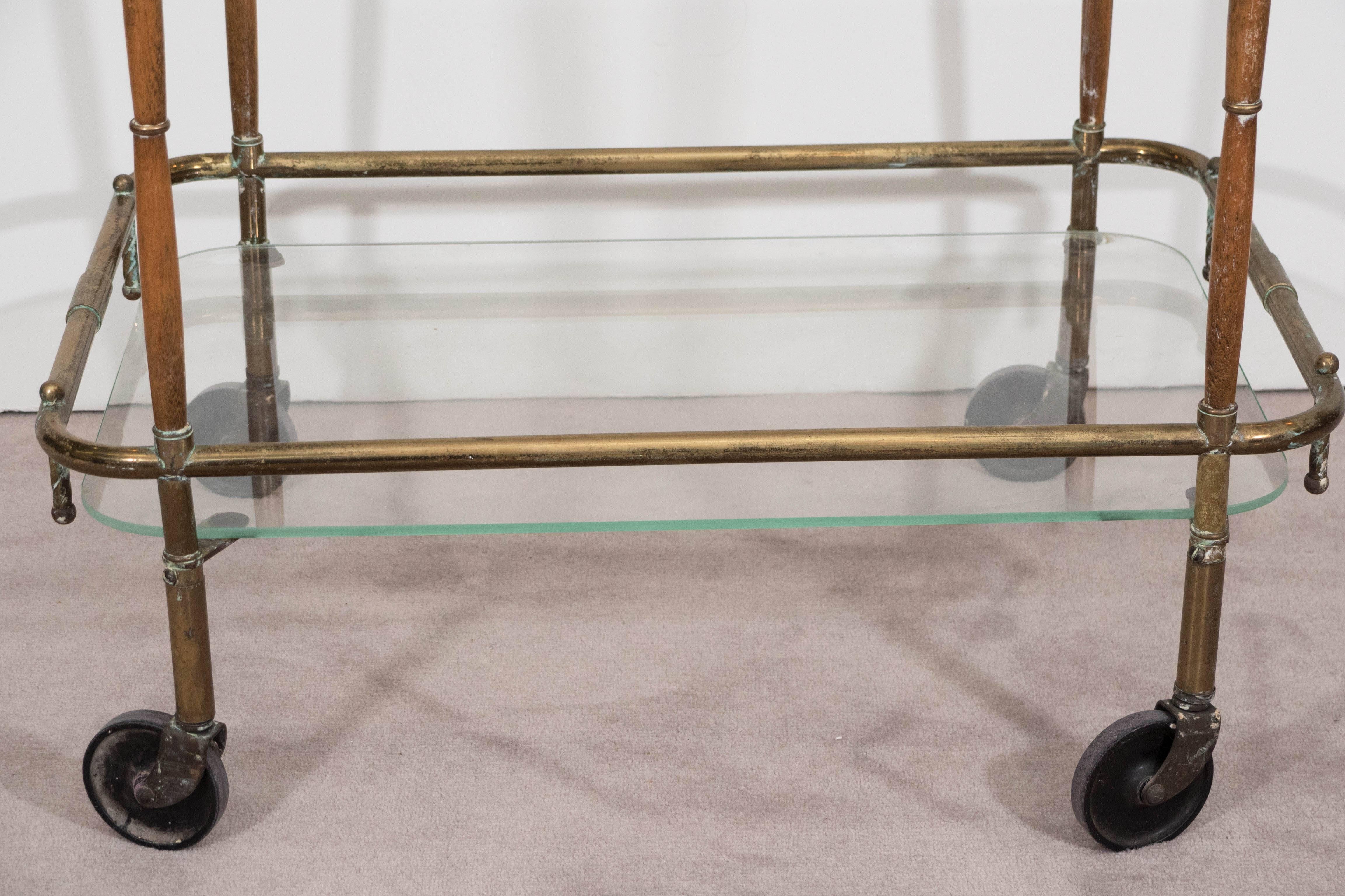 A bar cart and service trolley, produced in Italy circa 1960s, with two glass shelves against a tubular brass frame with push handle, accented with turned wood to the legs, on original casters. The cart remains in overall good vintage condition,