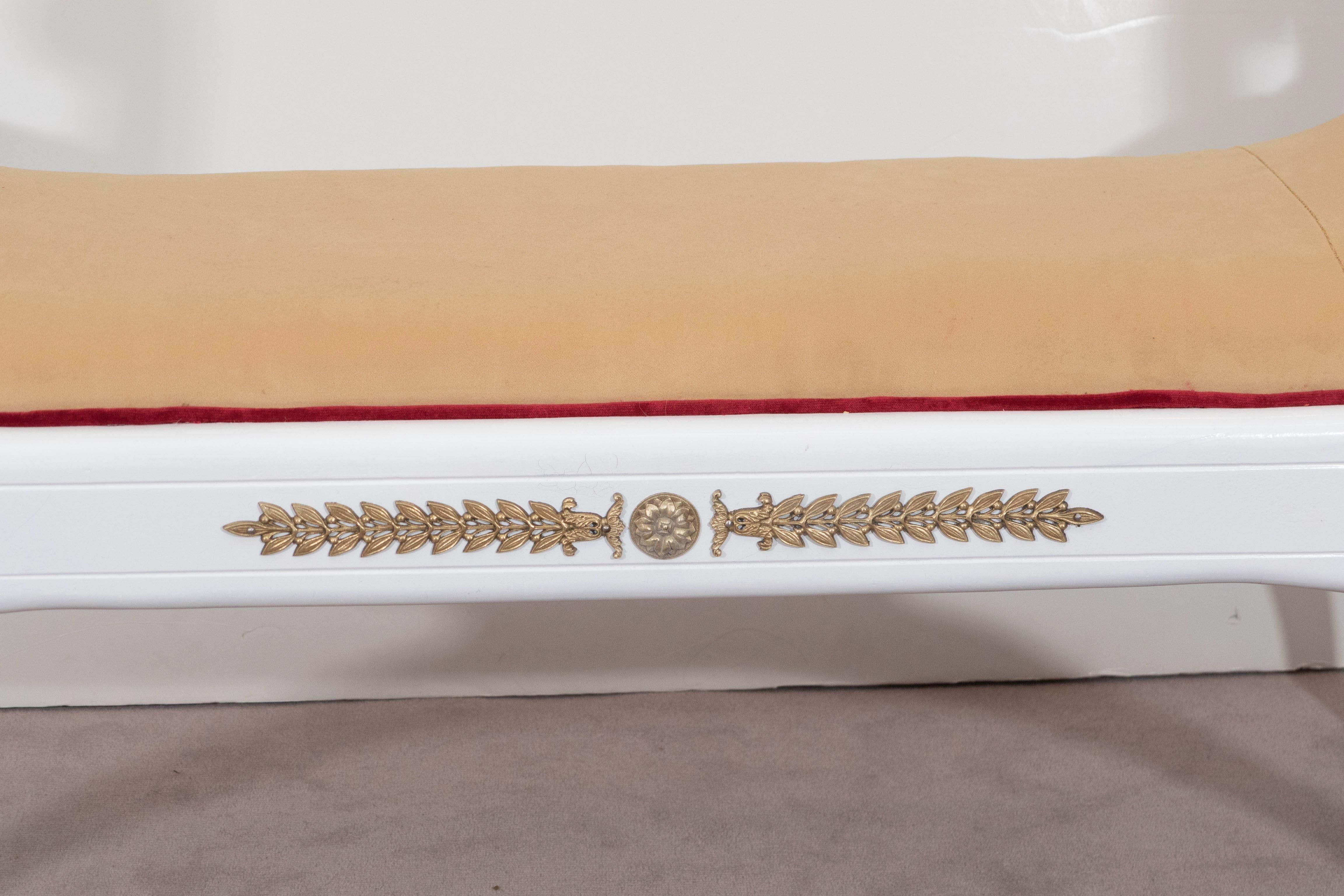 A window bench in the Regency style, with white painted finish, carved gilt lion's head uprights and turned arms, over cushioned seat, upholstered in velvet with red piping and Curule base, with bellflower and medallion mounts. The bench remains in