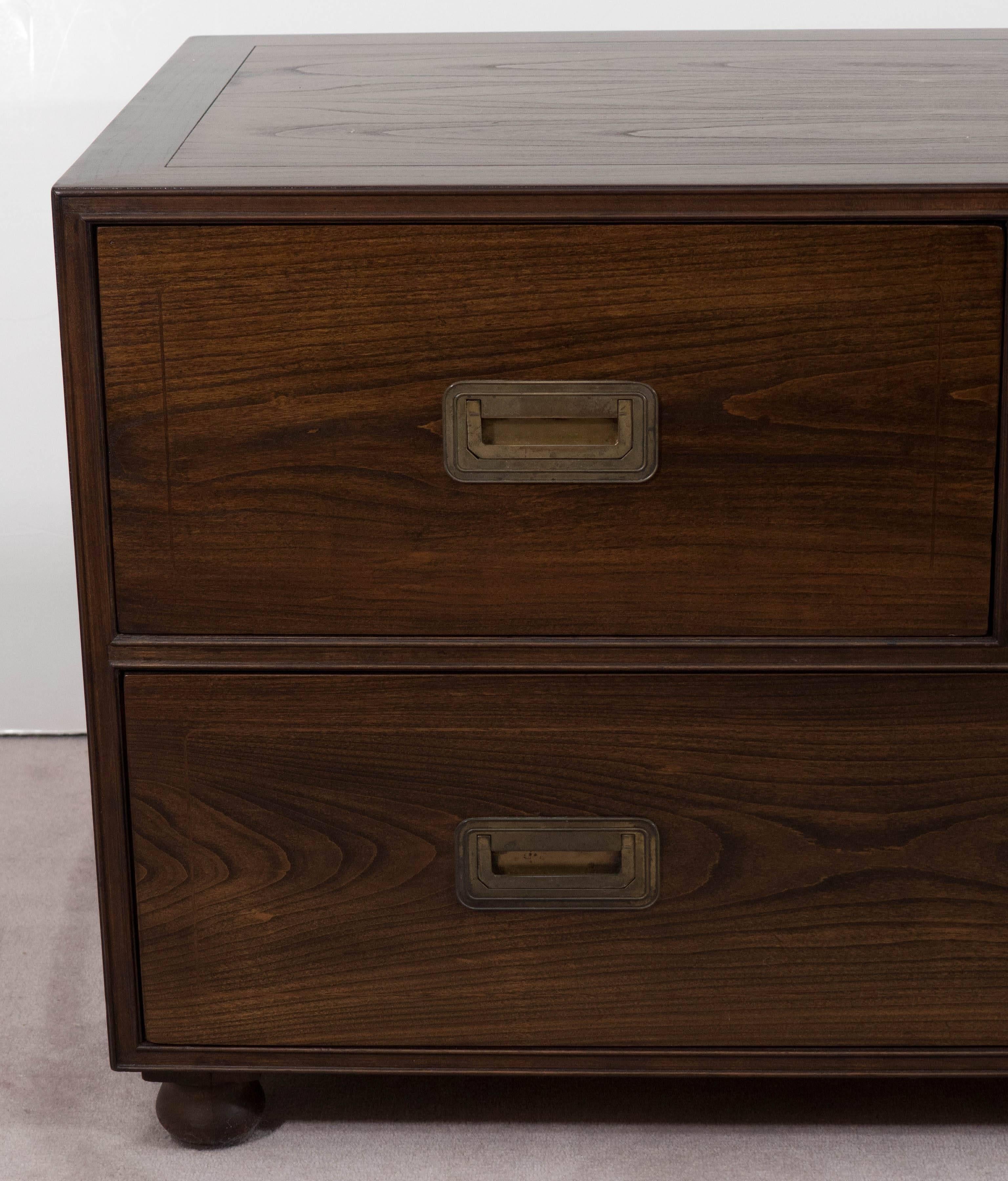 This rich toned wooden commode, produced circa 1960s-1970s by Baker Furniture, includes three drawers, two small and one lengthy, each with recessed pull handles in brass, on bun feet. Markings include the Baker Furniture plaque, inside the upper