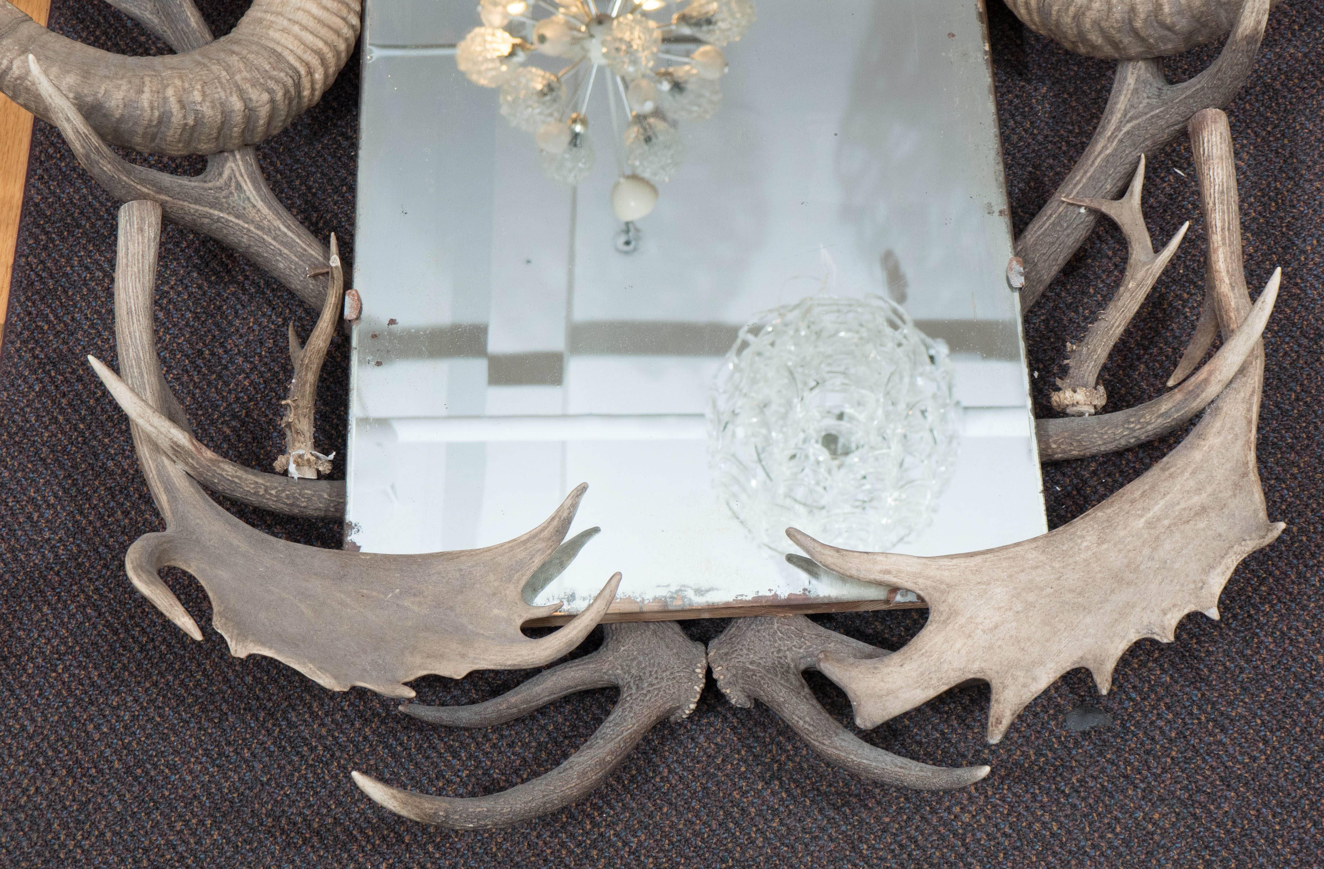 An early 20th century lengthy wall mirror, framed by an array of animal horns and antlers, affixed to a wood base, with rope suspension. Overall good vintage condition, with age appropriate wear to the hardware and slight warping, minor silvering