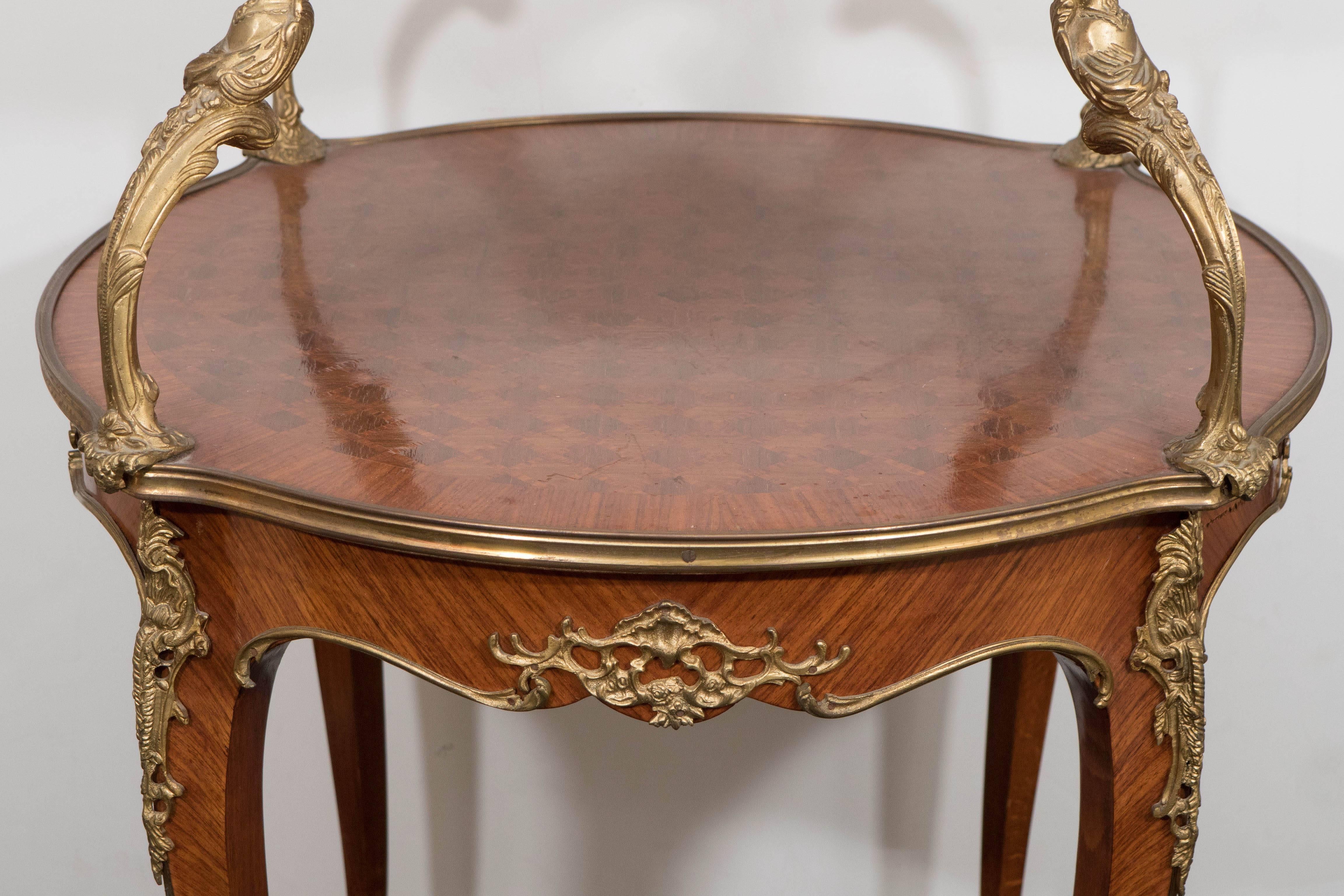 This Louis XV style serving table, produced within the early 20th century period, in the manner of designer and ébéniste François Linke, includes two round ogee edge tiers, each with kingwood parquetry tops, mounted with gilt bronze scrolling