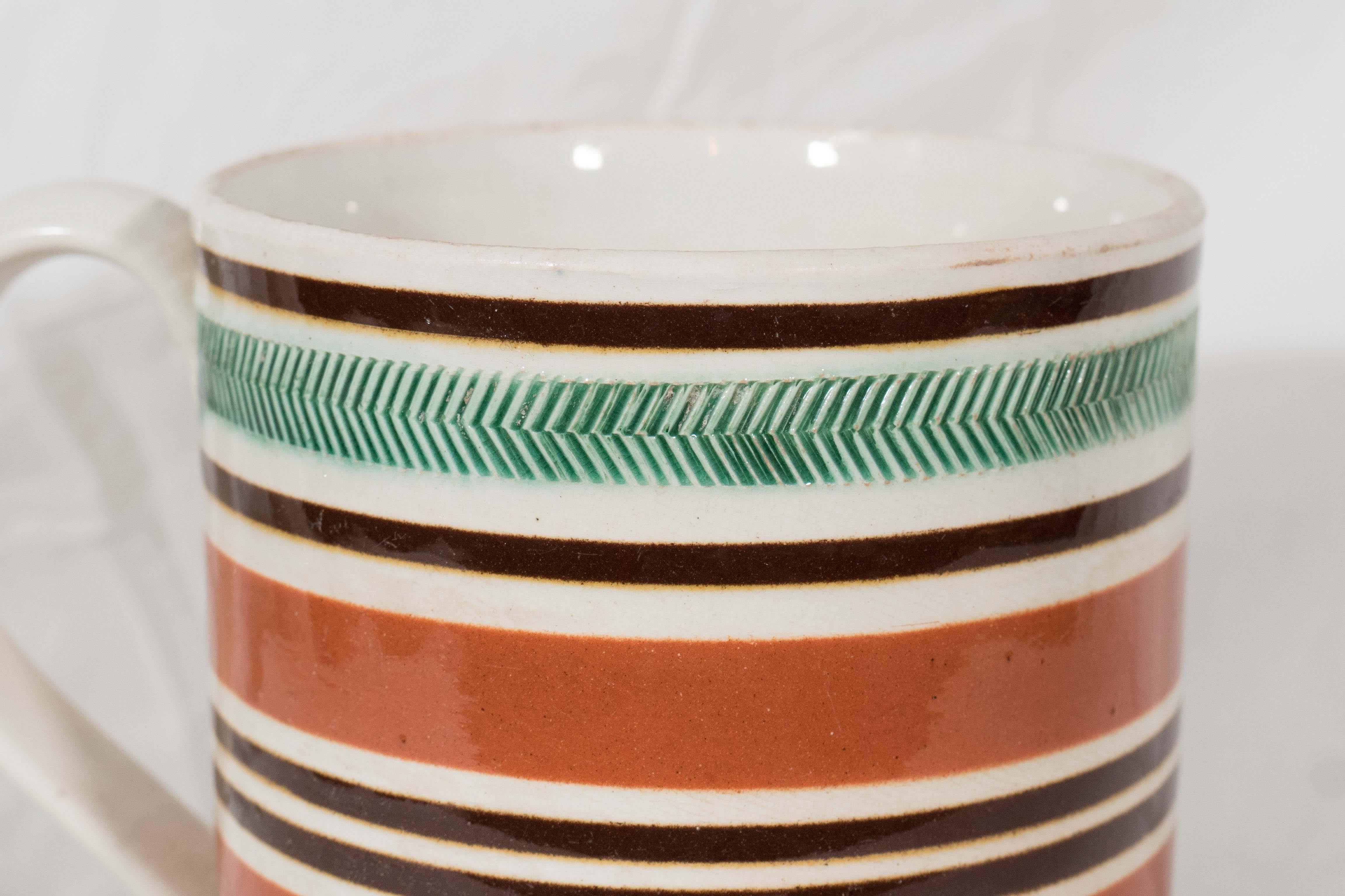 A large antique mocha mug with a pearlware body decorated with concentric bands of brown and green. Around the top of the mug is a band of incised rouletting with green glaze. Below that are alternating thin bands of dark brown and wider bands of