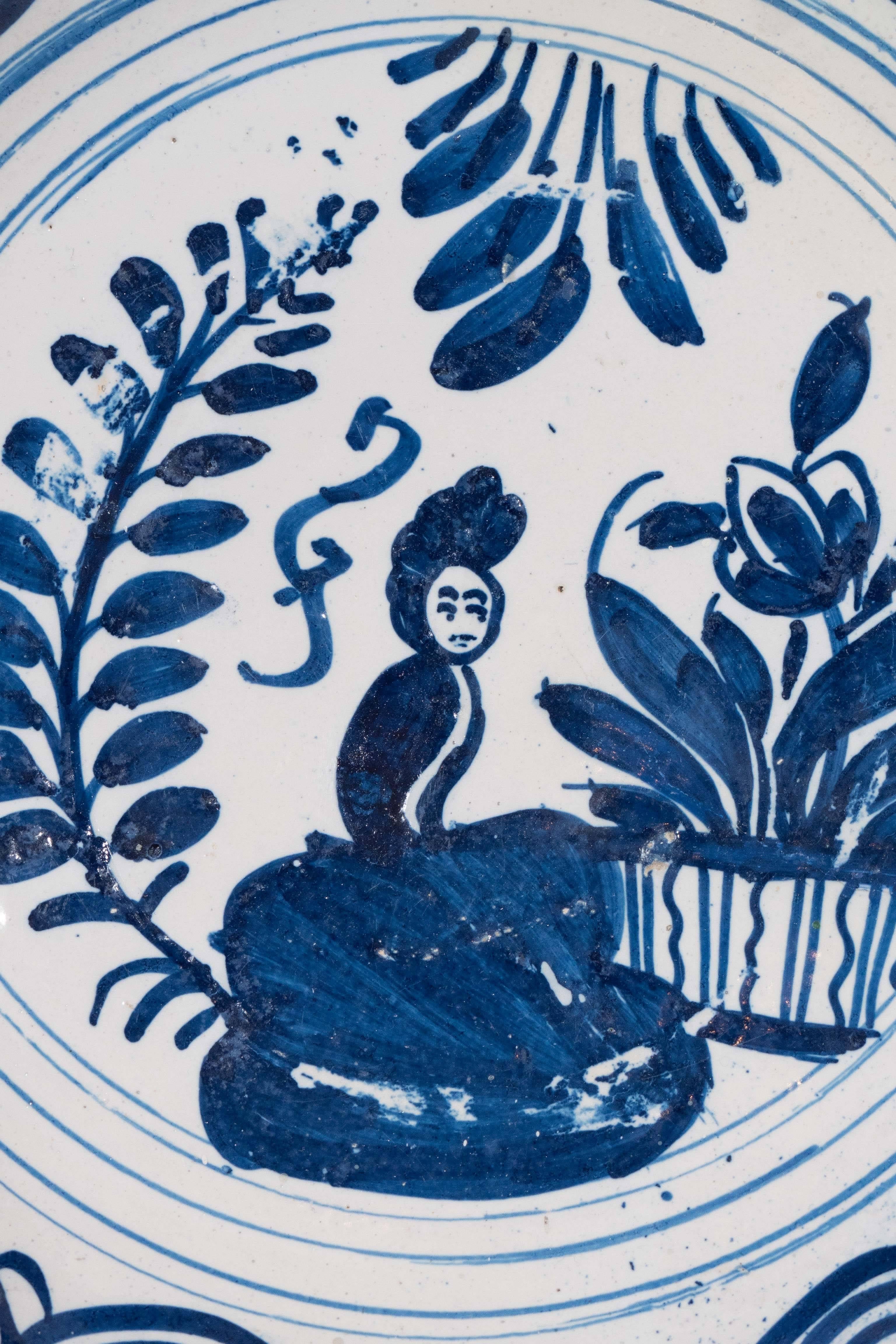 We are pleased to offer this early 18th-century Dutch Delft charger painted in cobalt blue with a scene showing a lady seated in a  garden. The border is decorated with linked concentric circles.
It is the naive aspect of this charger that makes it