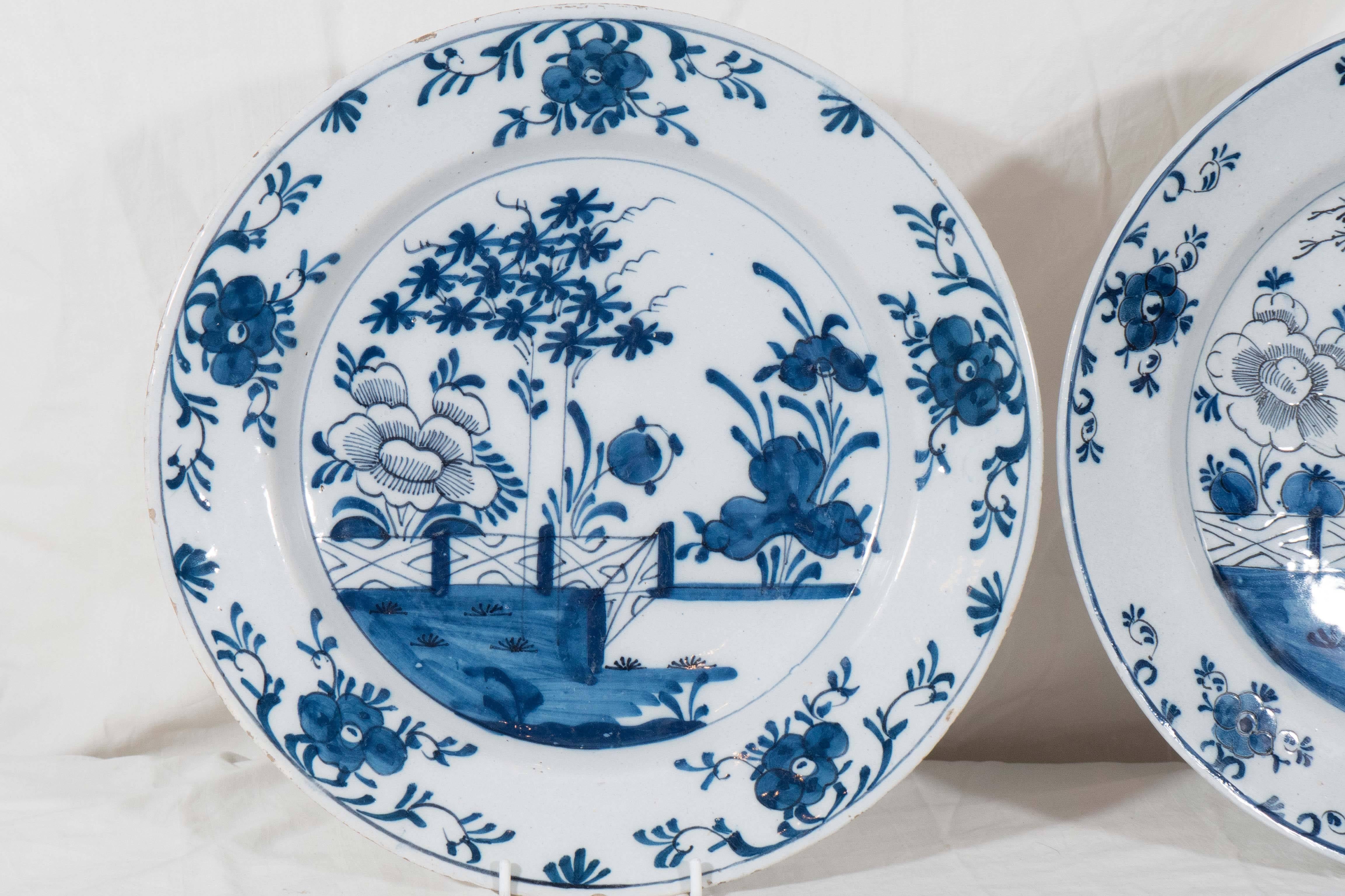 A pair of Dutch Delft chargers painted in cobalt blue showing a traditional chinoiserie garden with a very pretty flowering tree and an oversized peonie. The borders are decorated with scrolling vines and flower heads .