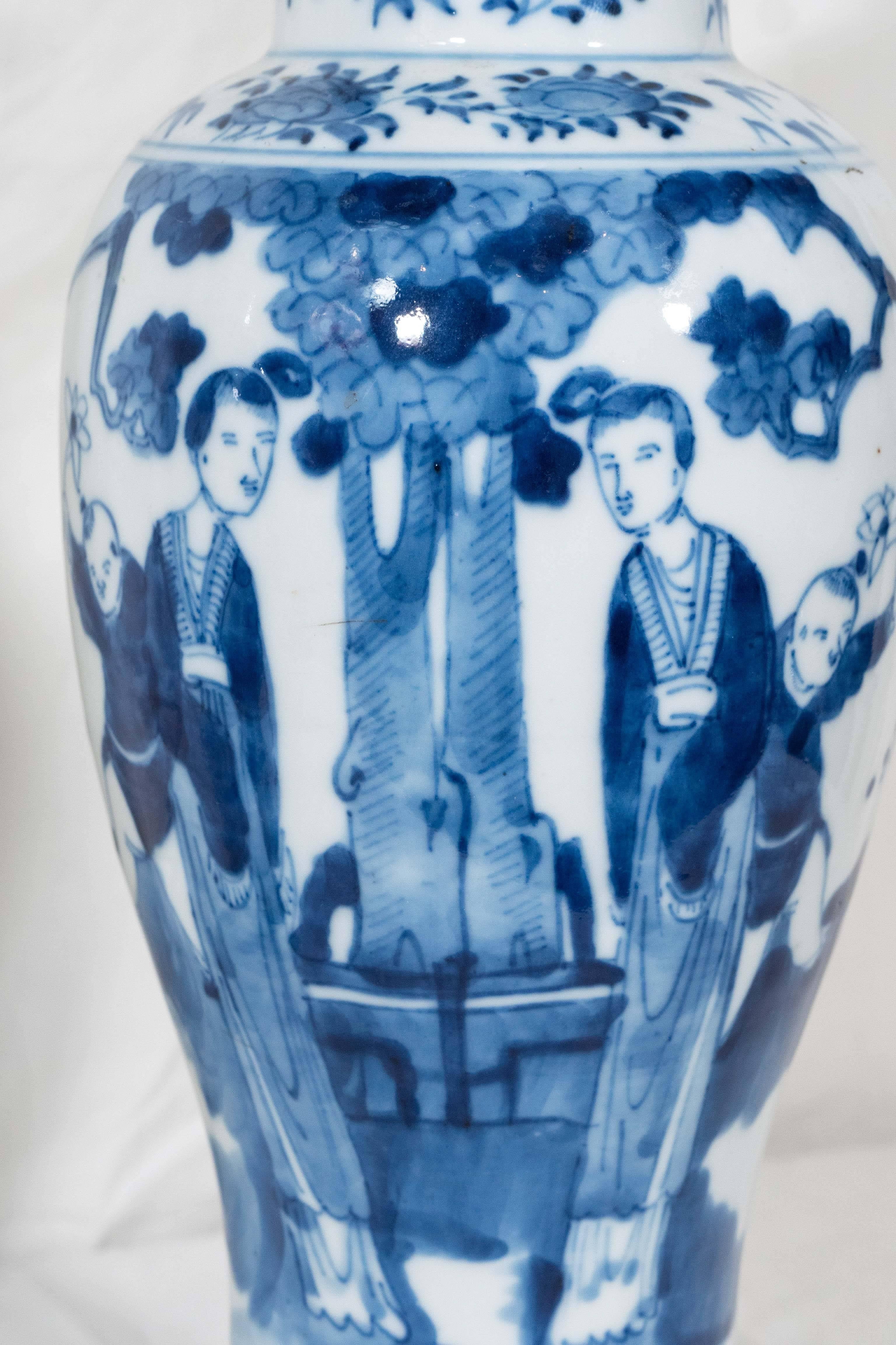 A pair of Chinese blue and white hand-painted porcelain vases decorated in underglaze cobalt blue showing a scene with a pair of Long Elizas and a young boy in a garden. Bands of flowers decorate the neck, cover, and base of each vase. The cover