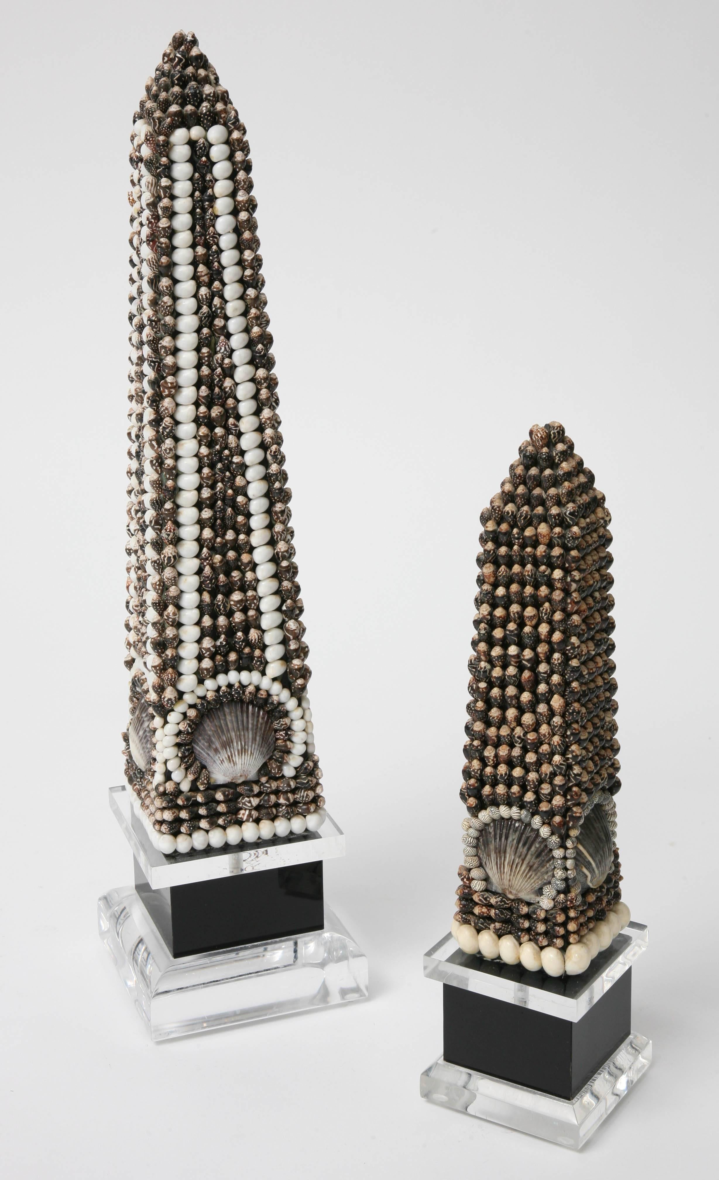 This set of obelisk are vey much in the style of pieces created by the iconic taste-maker Tony Duquette in the 1960s. They are fabricated of Lucite obelisk that have been encrusted with sea shells in dark bronze, black, beige and off-white
