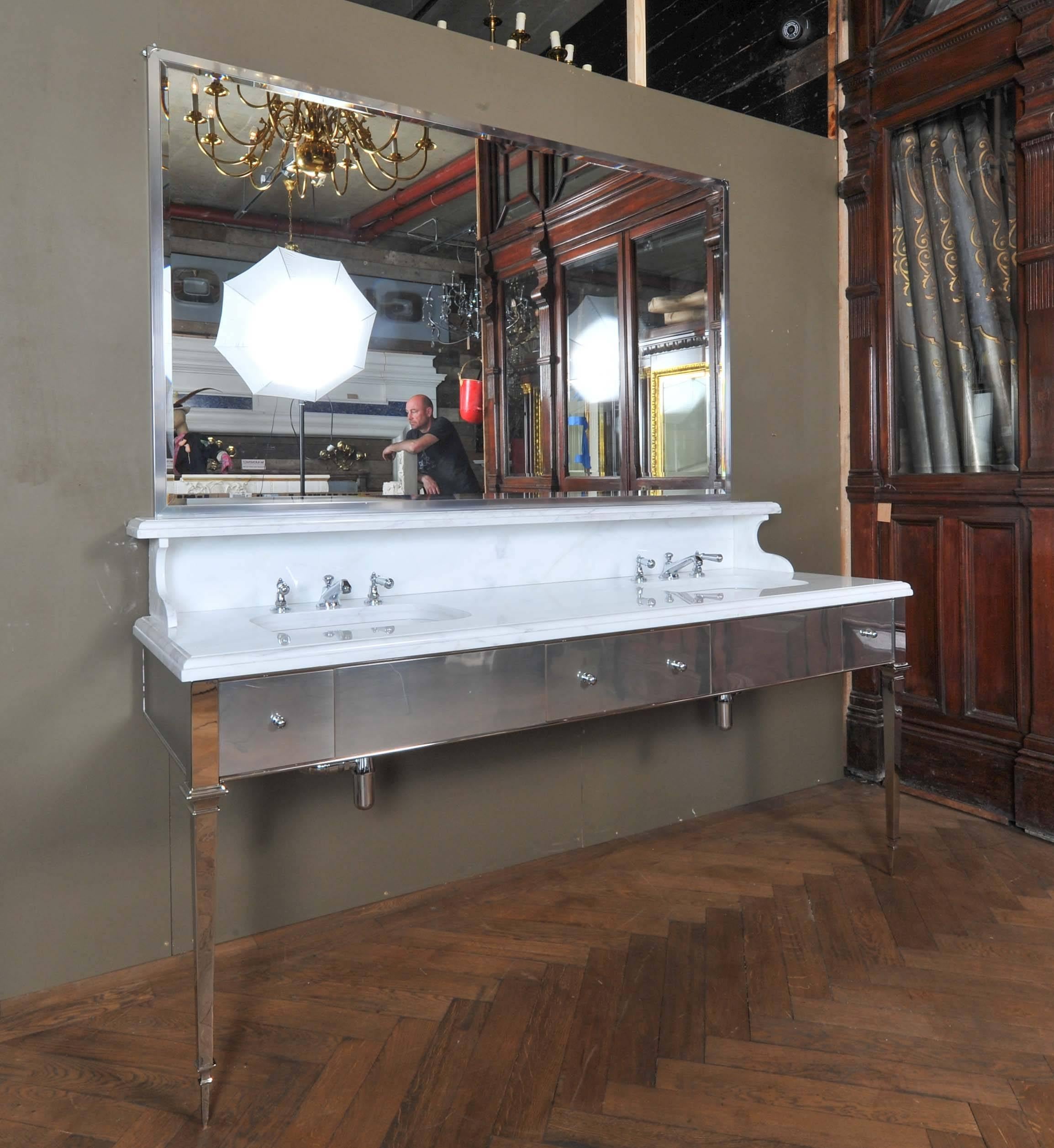 A deco style double sink washstand with a white Calcutta marble worktop, splashback and shelf and an elegant chrome base. The washstand feature polished chrome tapered legs and a brushed chrome front. The unit has a central soft-closing drawer, and