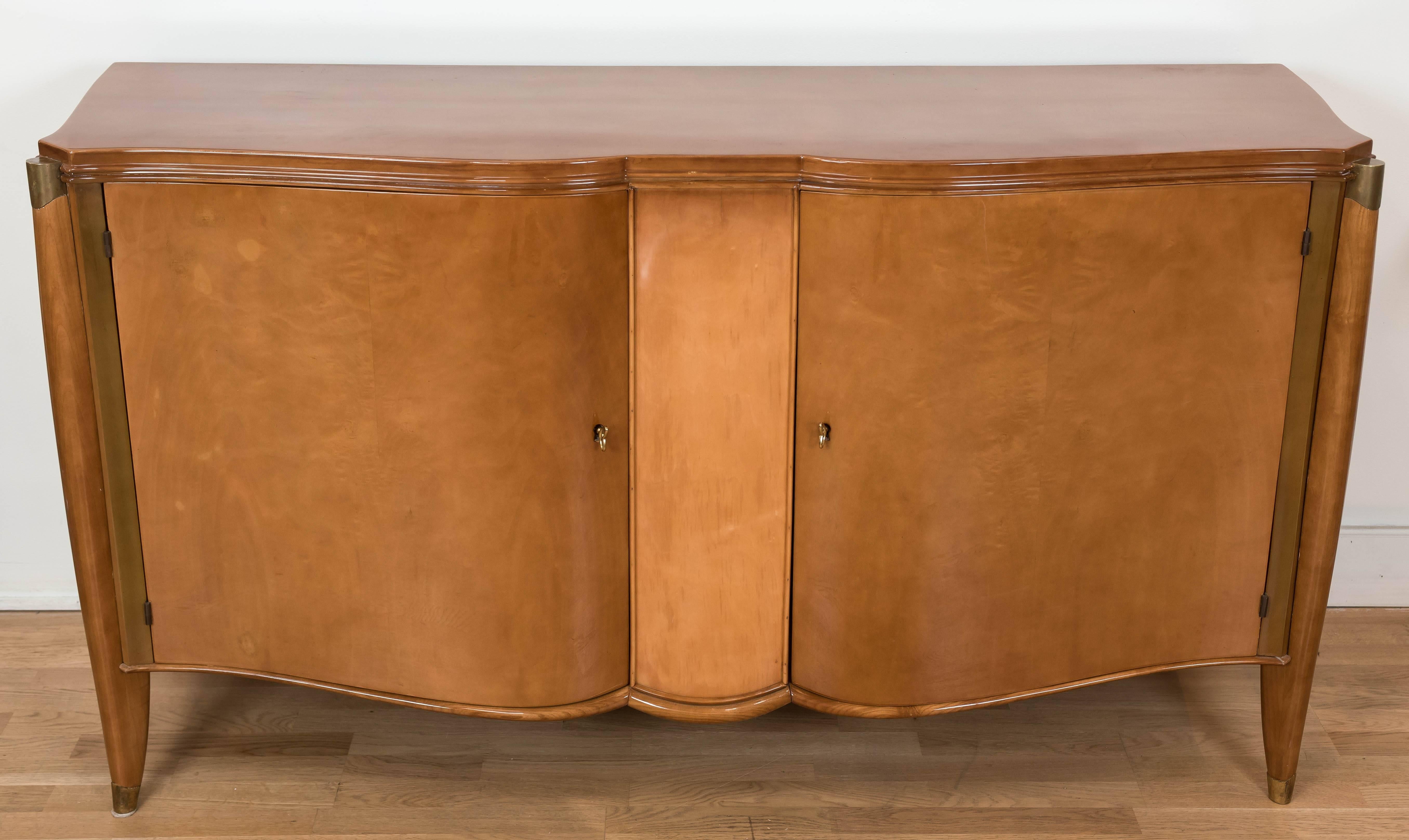 A  decadent and beautifully designed late Deco style serpentine front server with a blond oak interior and a rich maple veneered exterior. It is in its original high gloss lacquer finish and is accented with patinated brass details. The locks are in