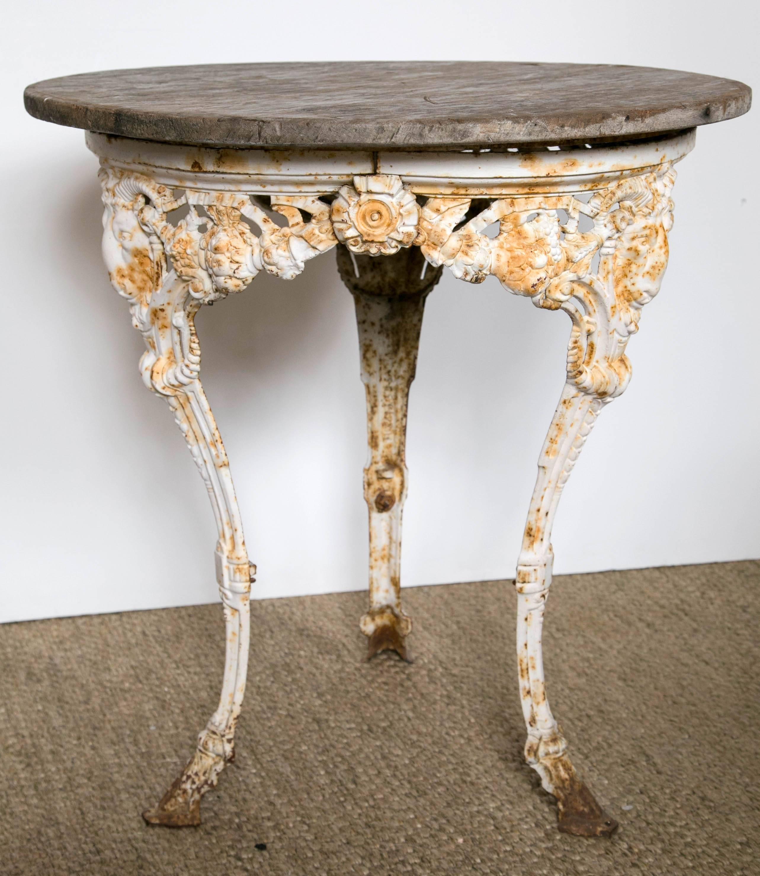 Vintage English pub table. Three cast iron legs feature decorative scroll work and ram heads. White finish and some patina on the iron. Wood top shows use appropriate wear.
