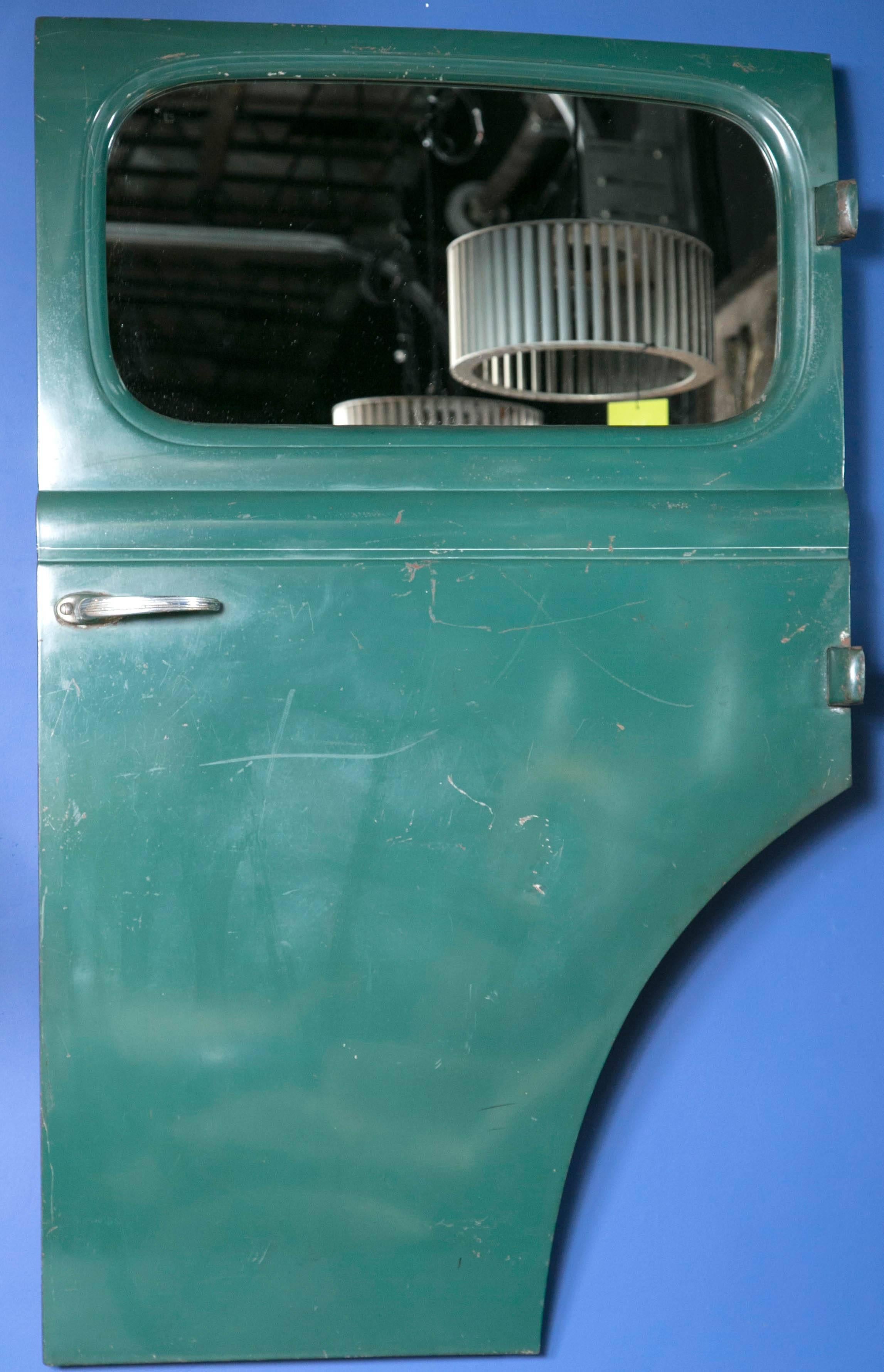 Dark green automobile rear door panel with the window removed and replaced with a mirror. The car door has been stripped on the inside and a mirror installed where the window was with wooden backing. The door has an exterior hinge and it was mounted
