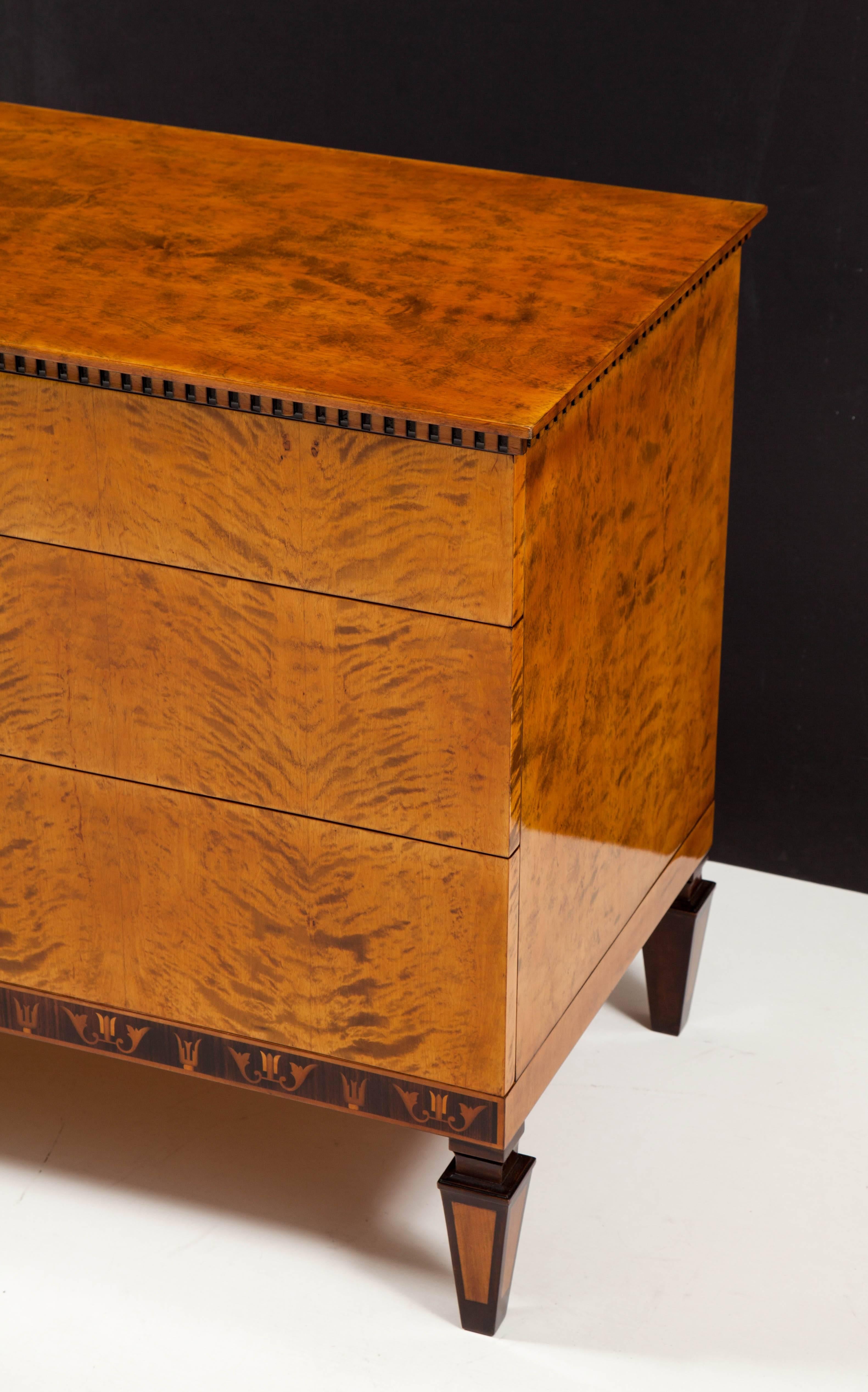 A Swedish grace flamed birch wood and palisander inlaid chest of drawers, circa 1930s, the rectangular top above a dentil molded frieze and three long drawers, the lower frieze with Classic Swedish Deco inlays raised on square tapered feet.
